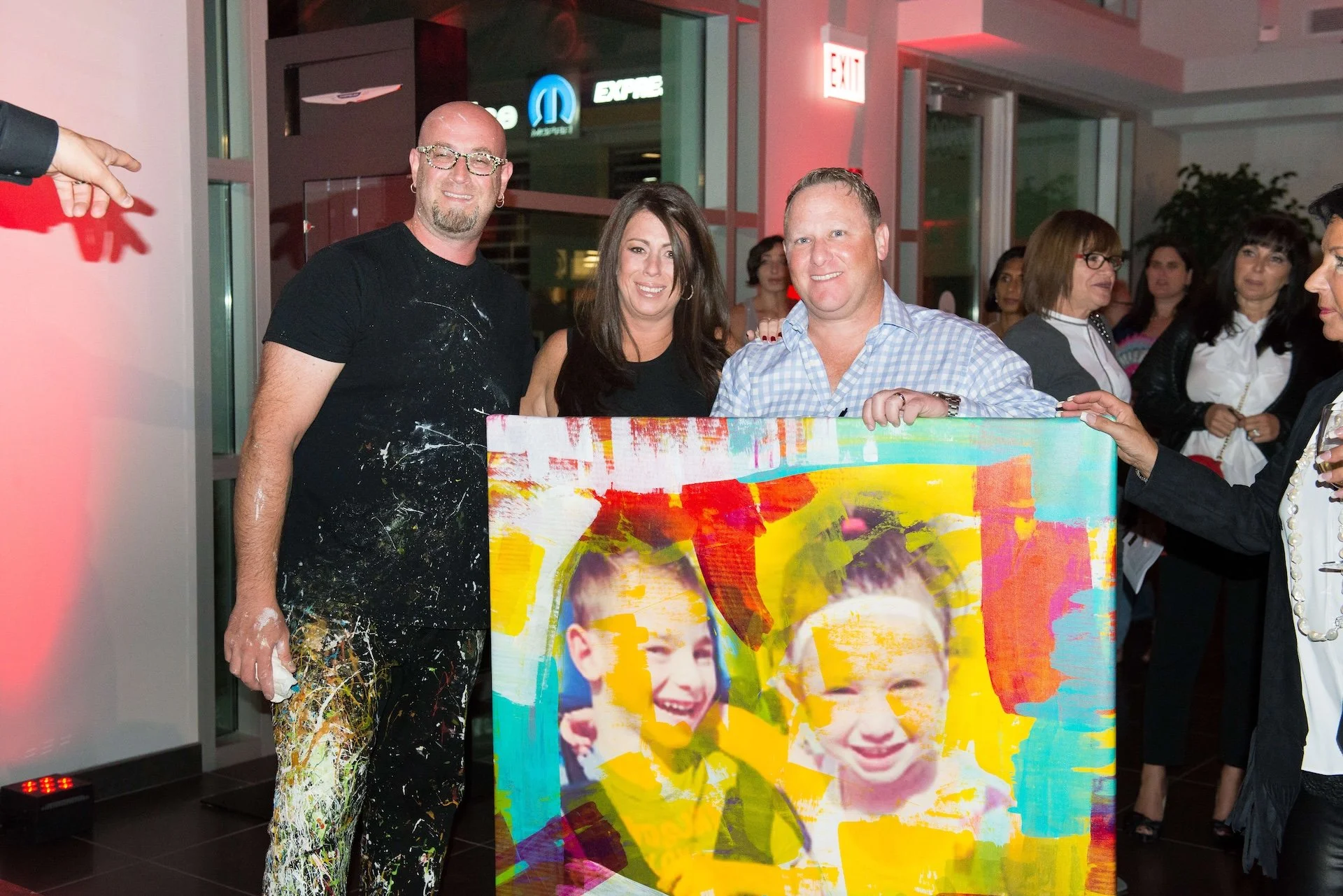 Three people smiling and standing behind a colorful, printed canvas with images of children. The person on the left is bald, wearing glasses, a black shirt with paint splatters, and paint-splattered pants. The person in the middle has dark hair, wearing a black sleeveless top. The person on the right has short blond hair, wearing a light blue checkered shirt. Several women are in the background, some wearing glasses and dark clothing, and others are engaged in conversation. The setting appears to be an art event or social gathering indoors near glass doors, with an exit sign and visible logo in the background.