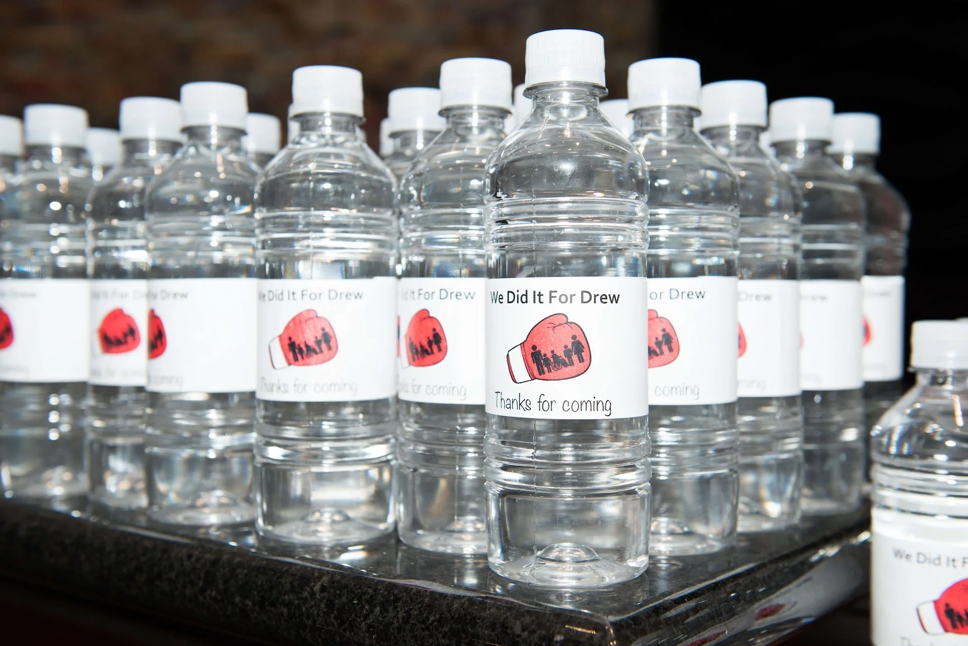 Multiple small plastic water bottles with white caps, arranged on a black surface, each with a personalized label featuring a pink heart and the message 'Thanks for coming'.