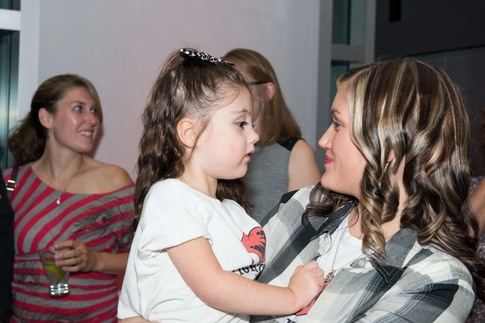 A woman and a young girl are facing each other closely, holding hands, and looking into each other's eyes in a warm interaction, with other women in the background.