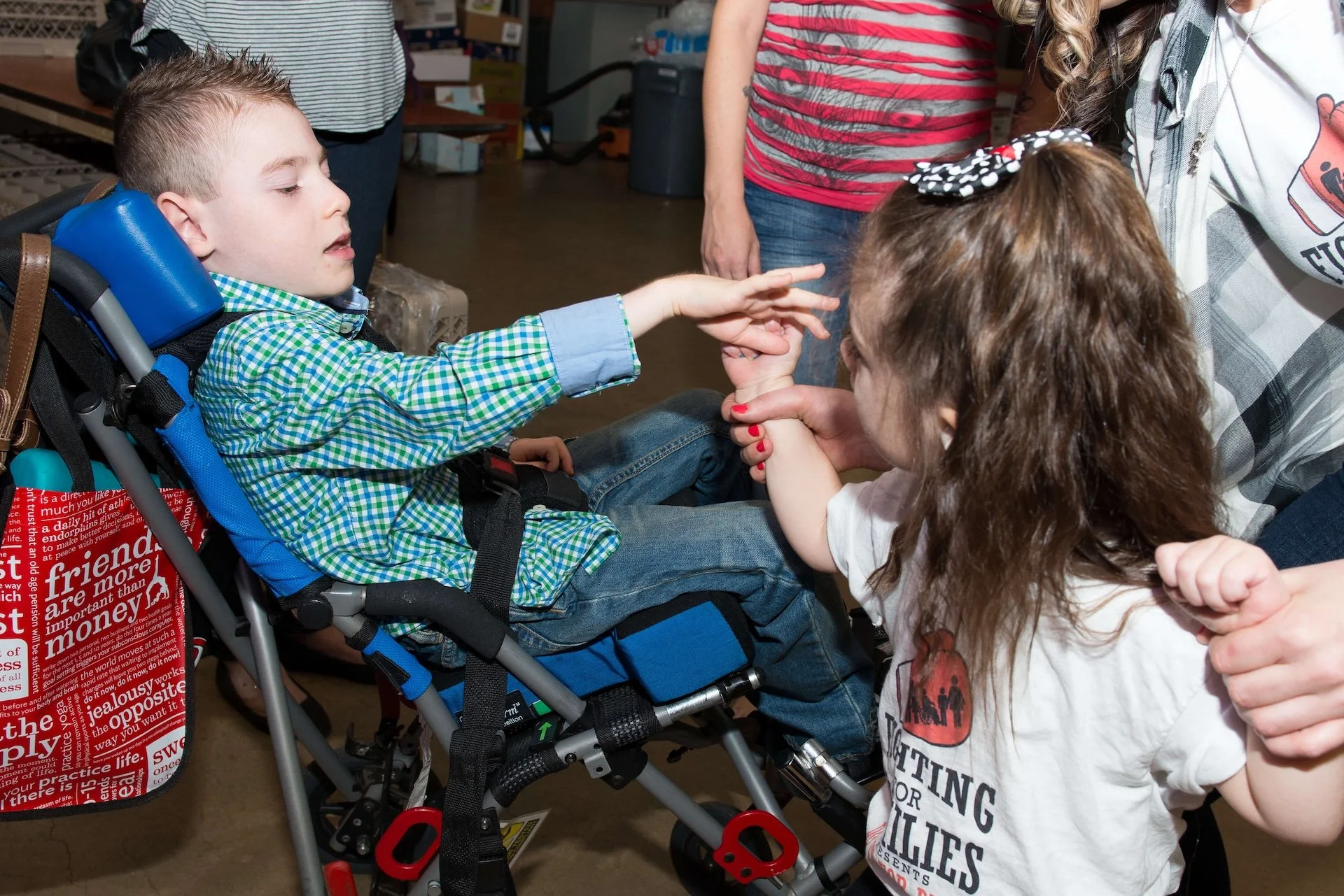 A young boy in a wheelchair reaching out and holding hands with a young girl, with two women standing nearby in a room filled with boxes and supplies.