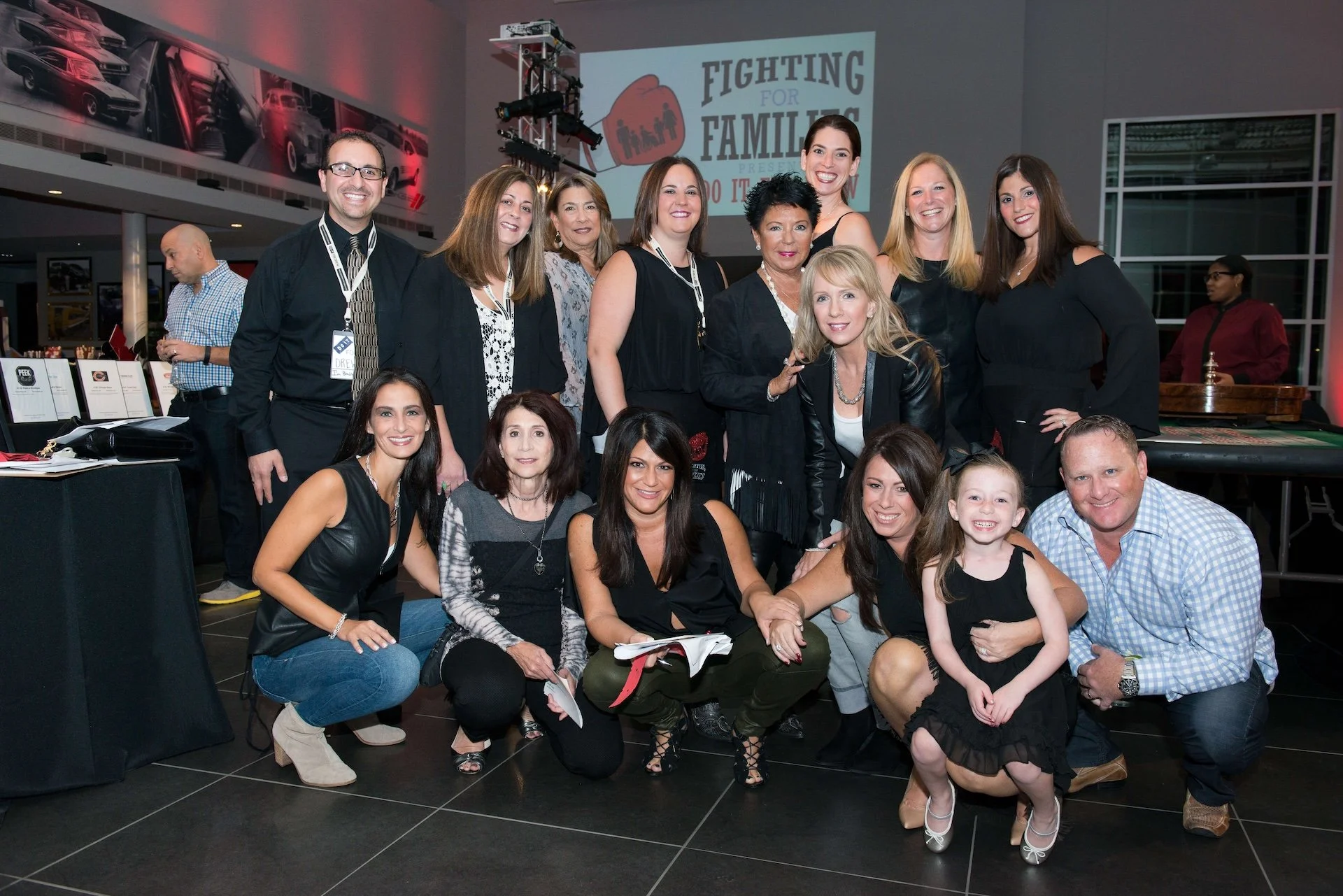 Group photo of diverse women and one man at a corporate event with a wall display and a screen showing 'Fighting for Families' in the background.