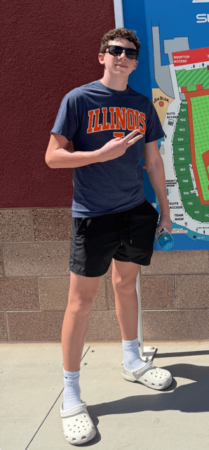 Young man wearing sunglasses, a navy T-shirt with 'Illinois' written on it, black shorts, white socks, and Crocs, standing outdoors near a stadium seating chart.
