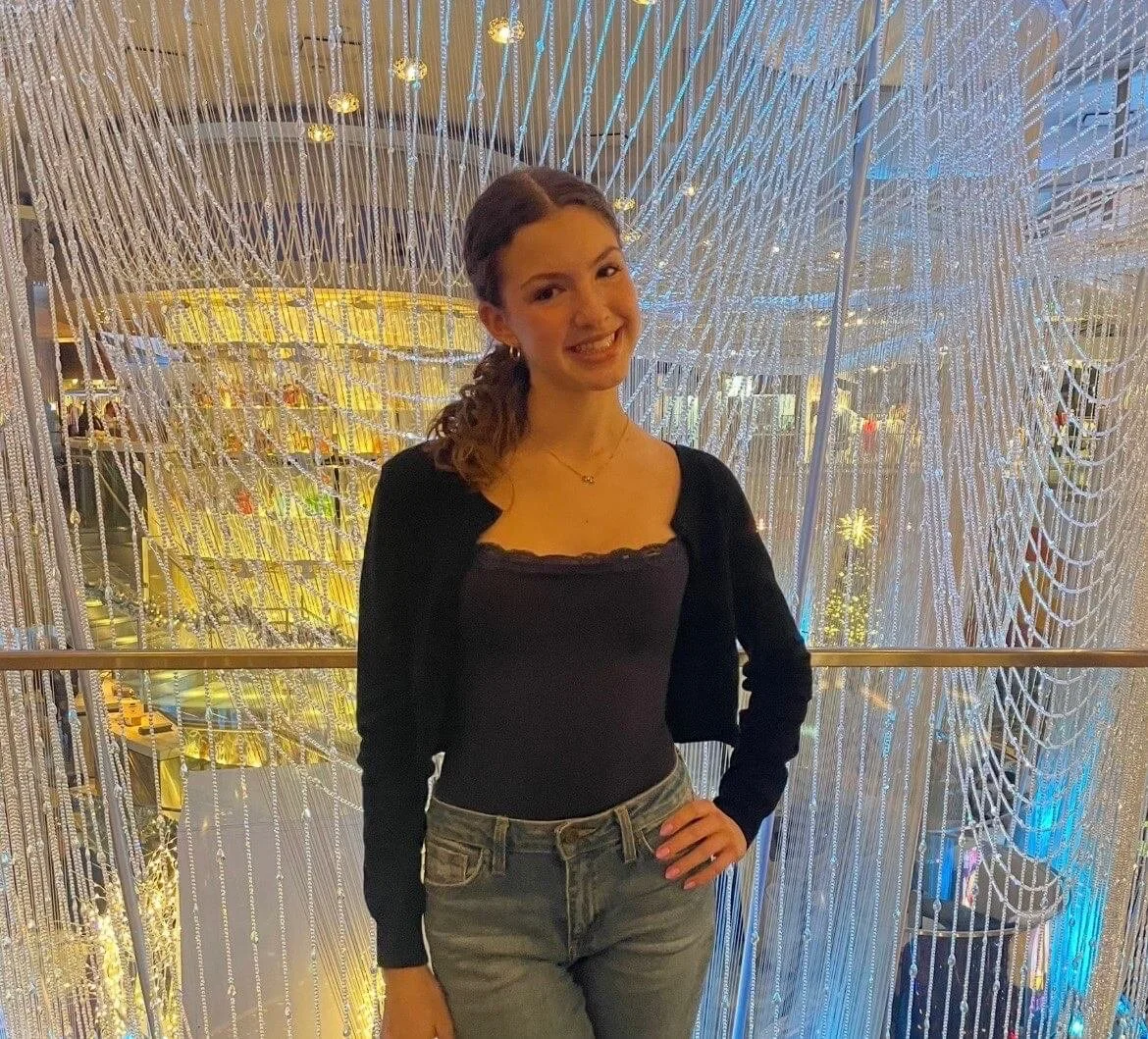 Young woman standing in front of a large decorative light display made of hanging strings of lights, smiling and wearing a black top with a black cardigan and gray jeans.