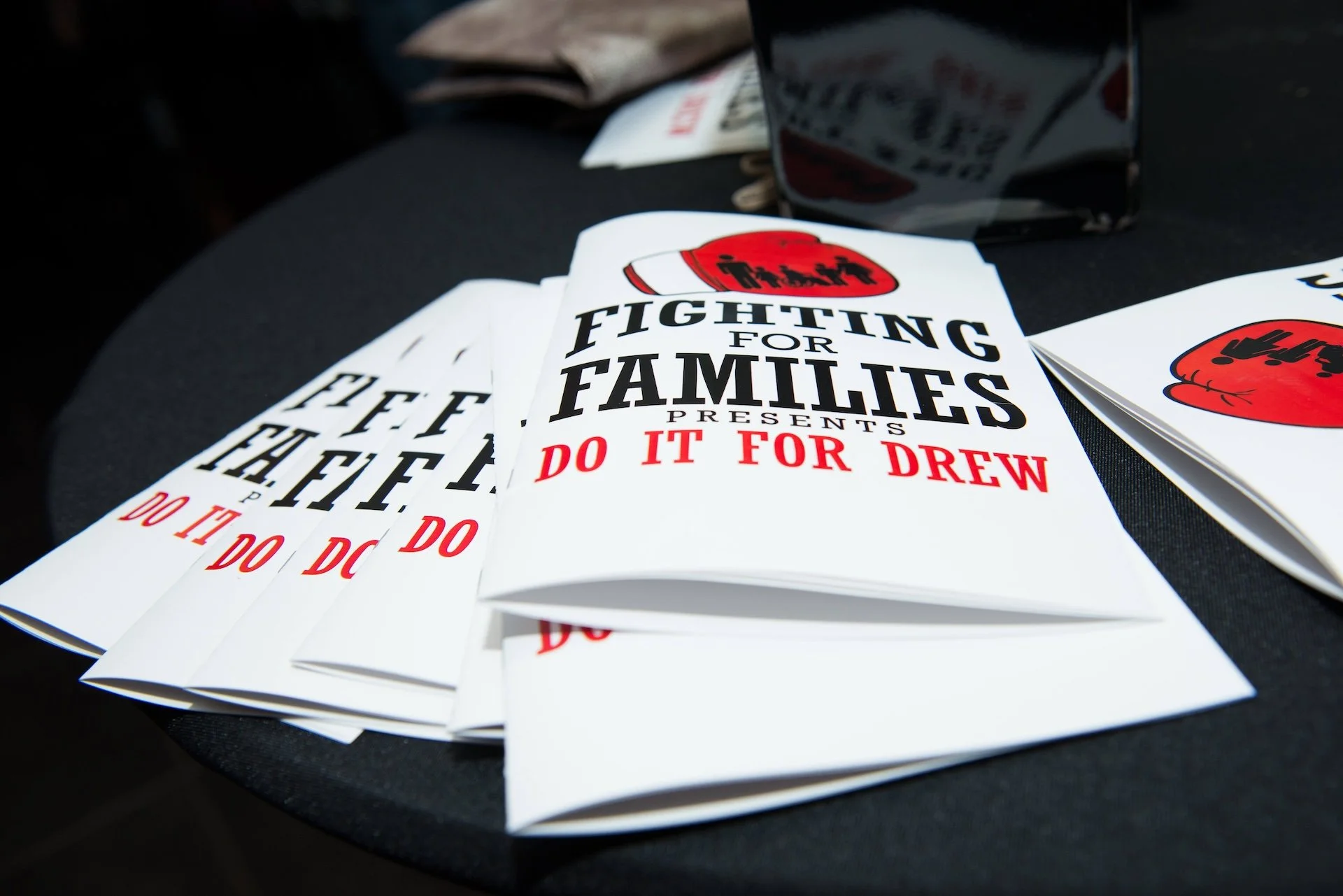 Flyers on a black table advocating fighting for families, with bold text saying "FIGHTING FOR FAMILIES" and "DO IT FOR DREW" in red.