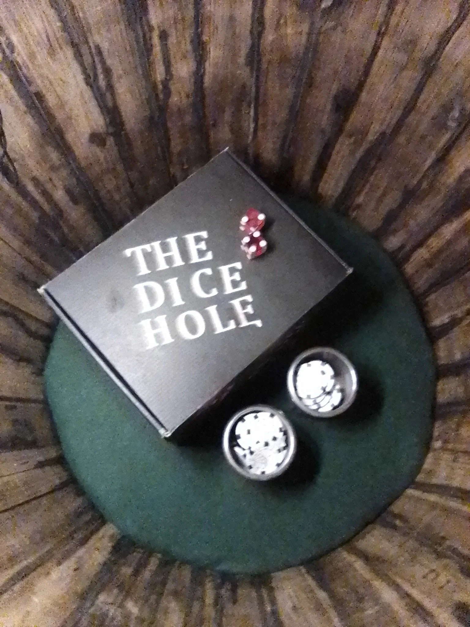 A black game box labeled 'The Dice Hole' with two red dice on top and two bowls filled with white poker chips, all placed on a round green surface against a wooden wall background.