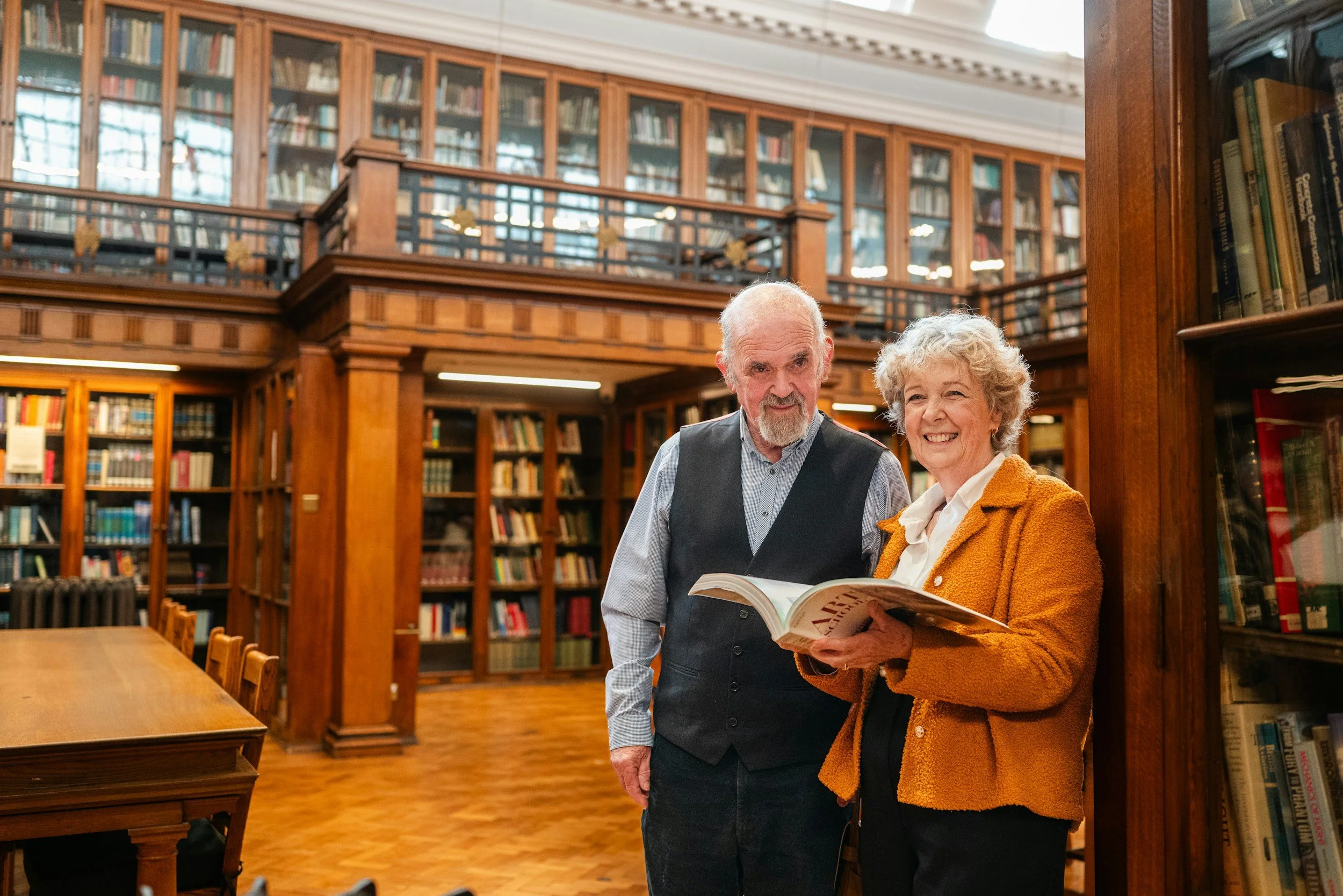 Two elderly people, a man and a woman, standing in a library and looking at a book together, surrounded by wooden shelves filled with books.
