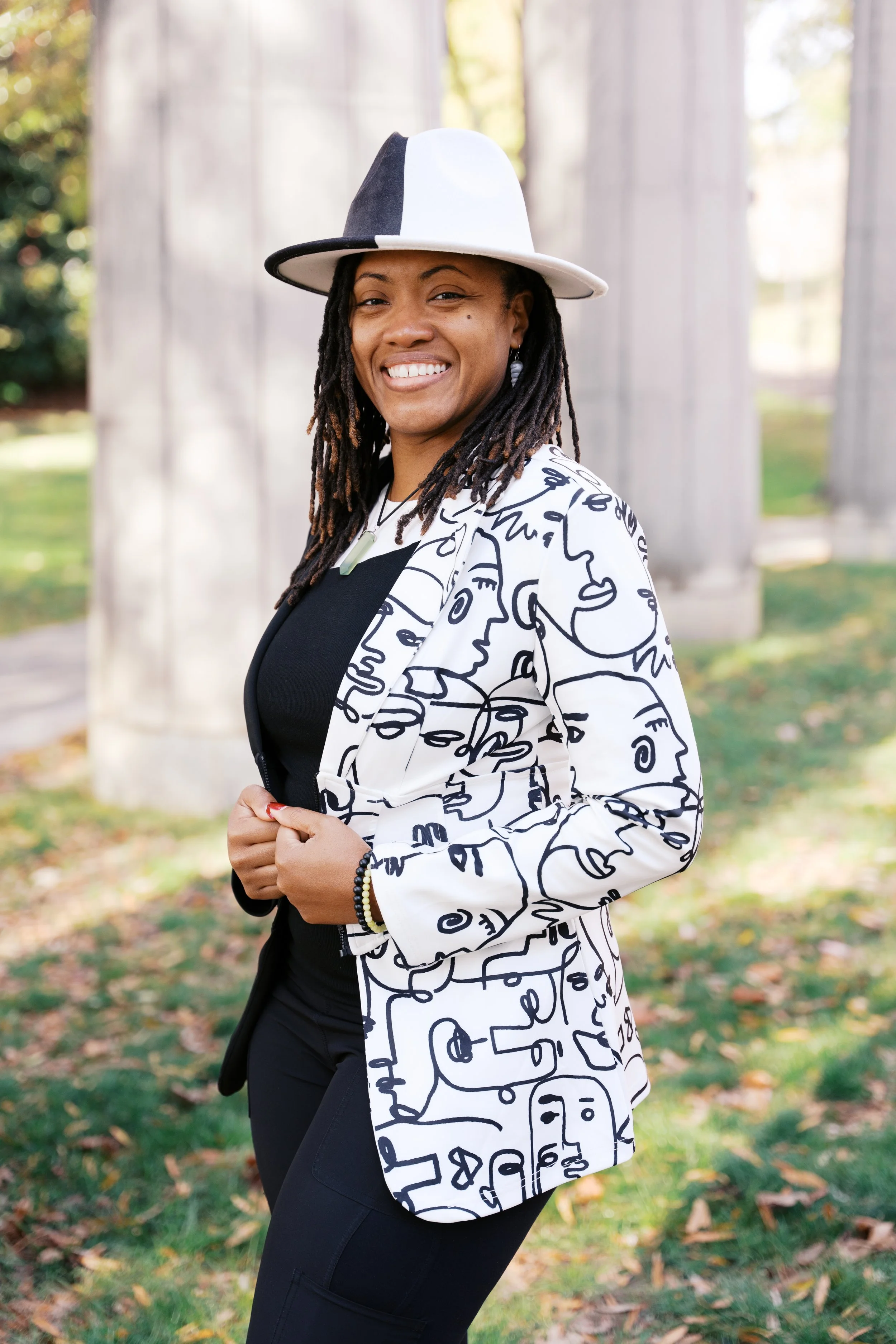 A woman smiling outdoors wearing a black and white abstract patterned blazer and a white and black wide-brimmed hat, with a background of green grass, fallen leaves, and trees.
