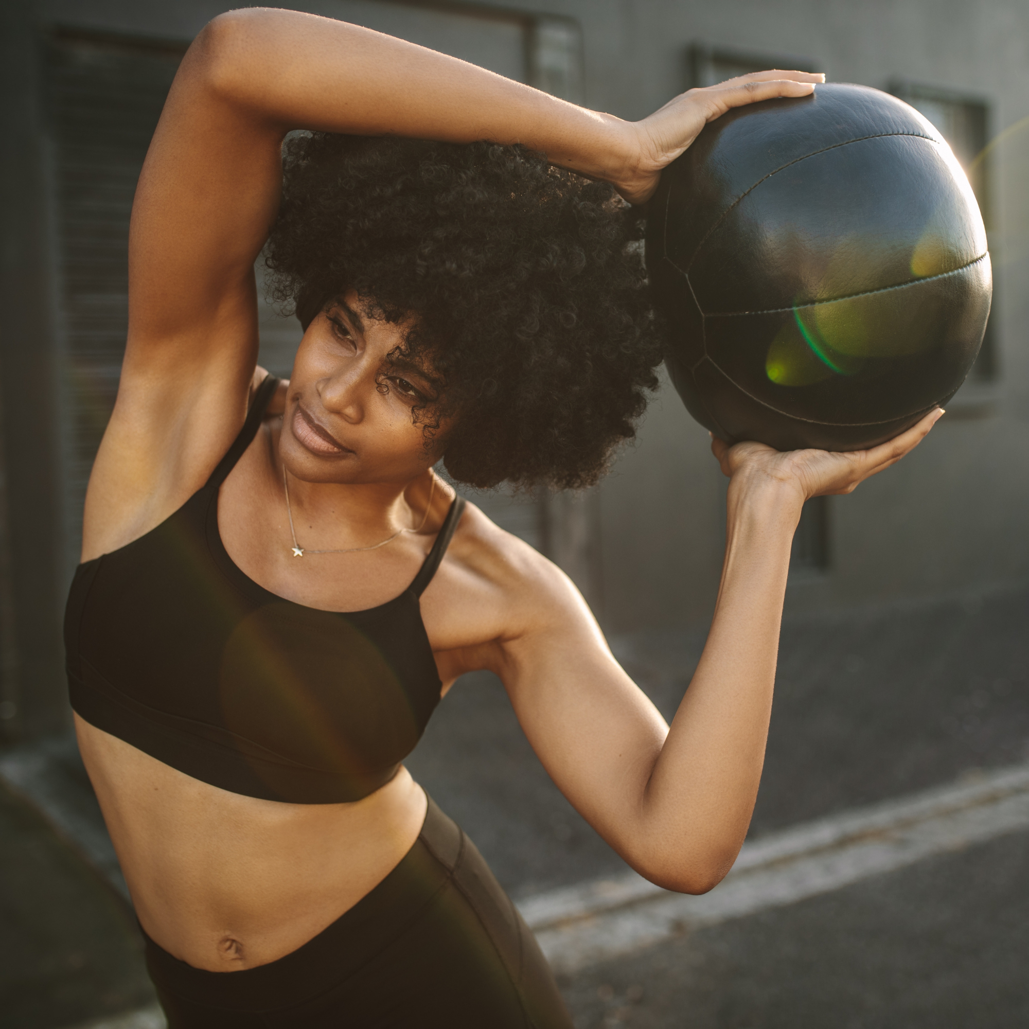 A woman with curly black hair lifting a black medicine ball while doing a workout outdoors.
