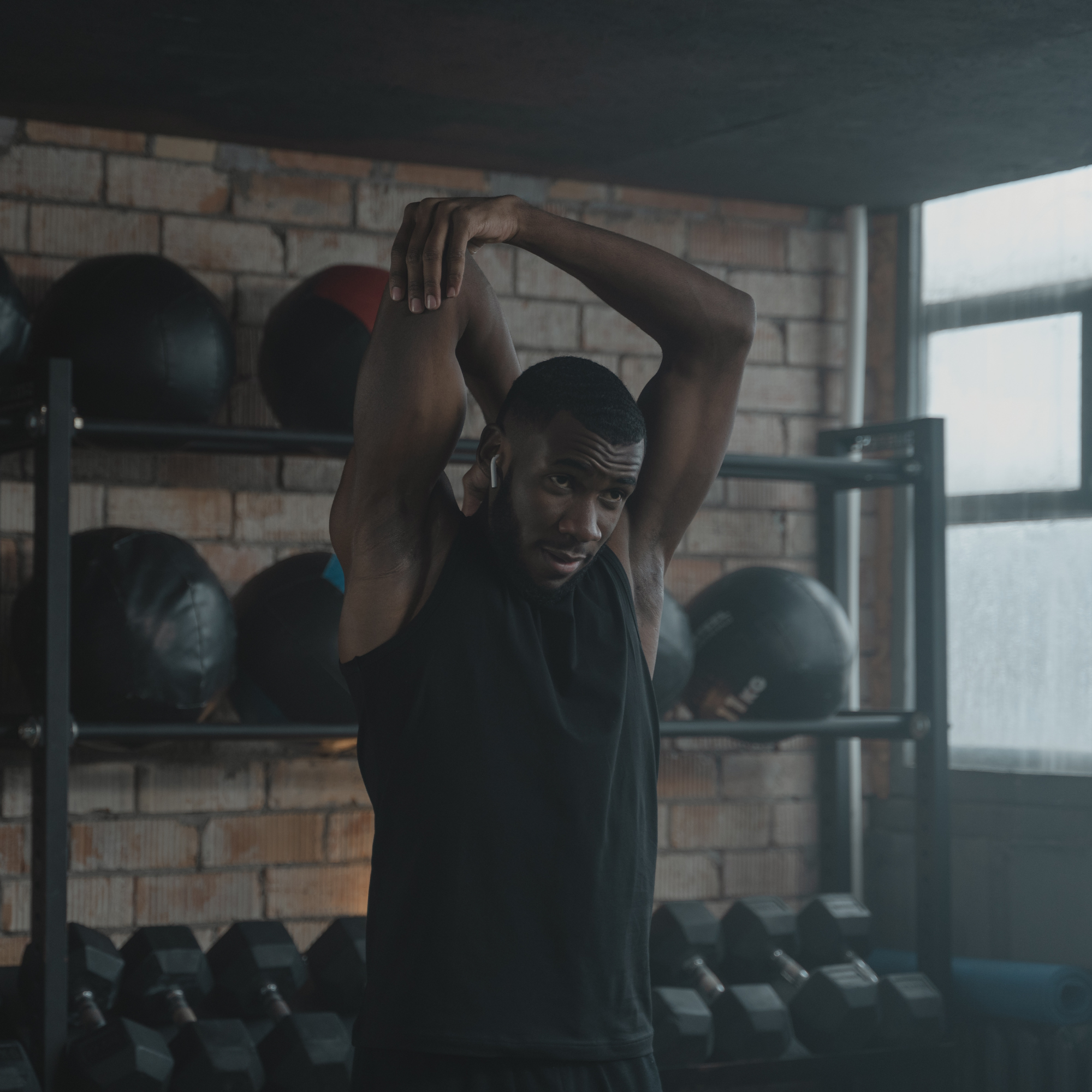 A man with dark skin, short curly hair, wearing a black shirt, is in a gym lifting or stretching his arms above his head. The gym has a brick wall, weight racks, and a shelf with medicine balls in the background.