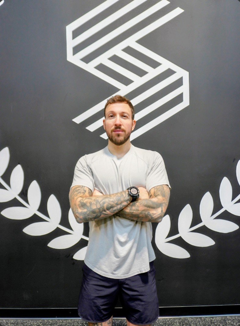 A man with tattoos on his arms, wearing a white athletic t-shirt and black shorts, standing with arms crossed in front of a dark wall with a geometric logo and white laurel wreath design.