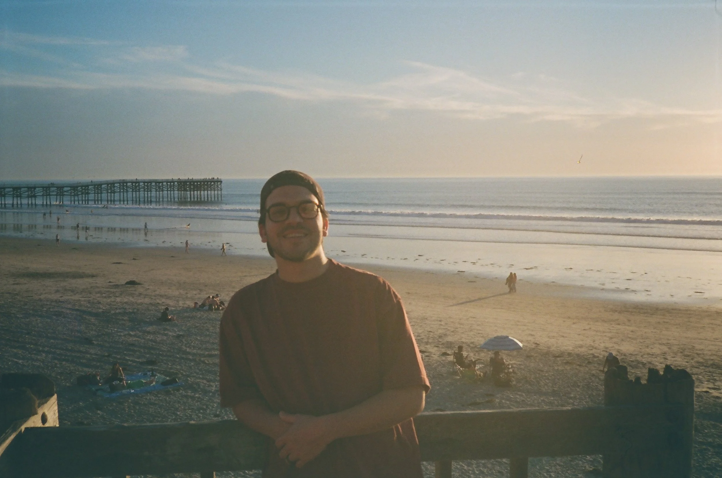A man wearing glasses and a backward cap standing on a beach during sunset, with people in the background, a pier extending into the ocean, and a seagull flying overhead.