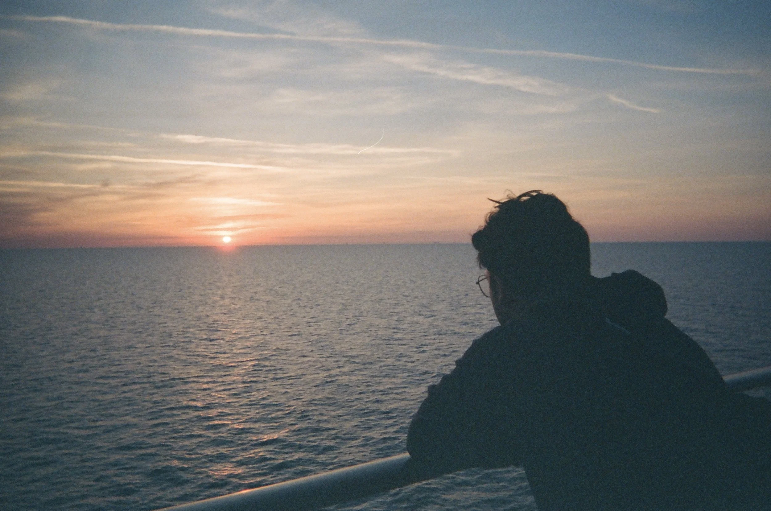 A person with dark hair wearing glasses and a dark jacket is sitting on a boat or dock, gazing at a sunset over the ocean. The sky is partly cloudy with a crescent moon visible, and the sun is near the horizon, casting a warm glow on the water.