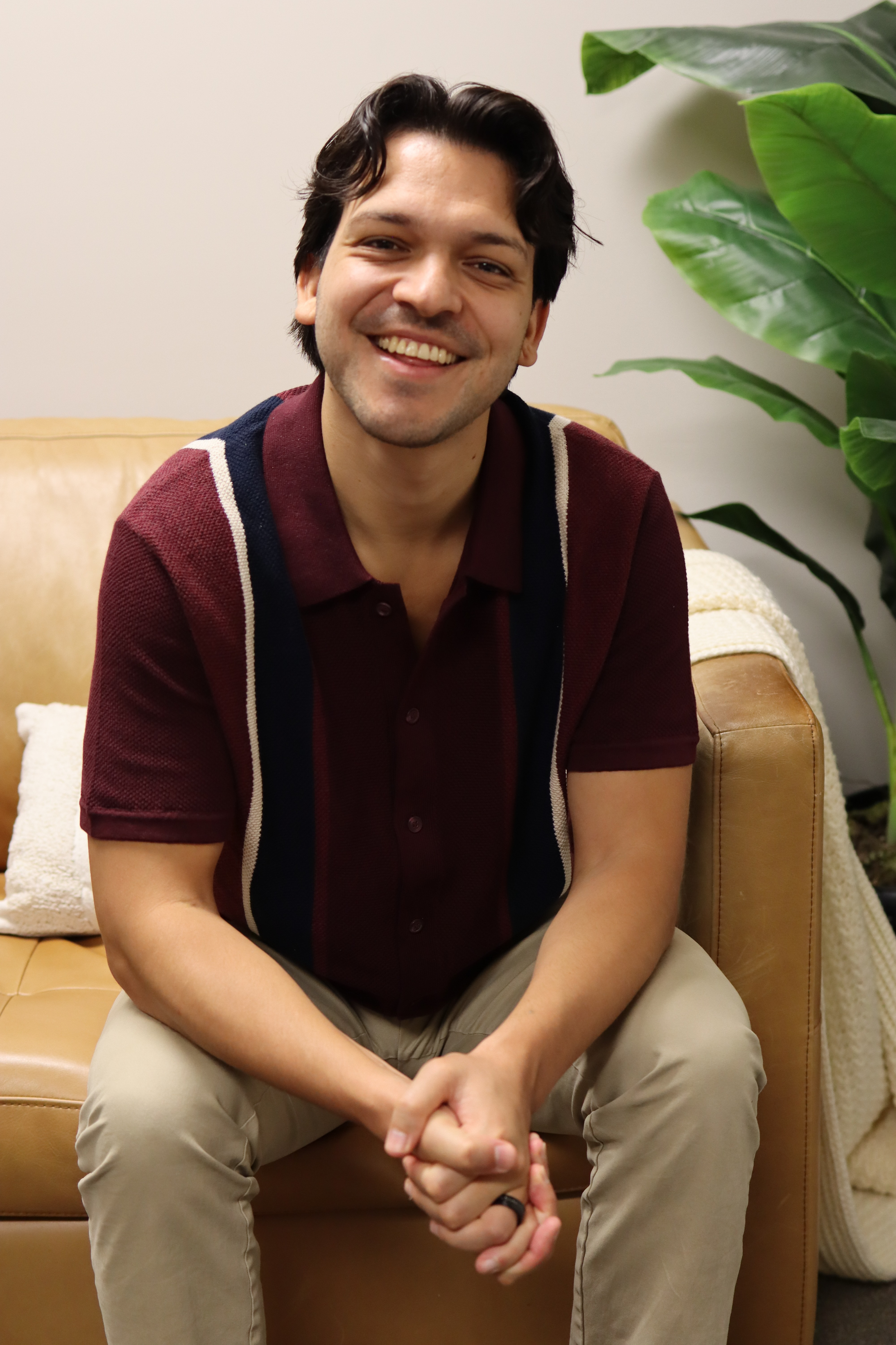 Smiling young man with dark wavy hair sitting on a tan leather sofa, wearing a maroon polo shirt, beige pants, and a dark ring on his left hand, with green leafy houseplant in background.
