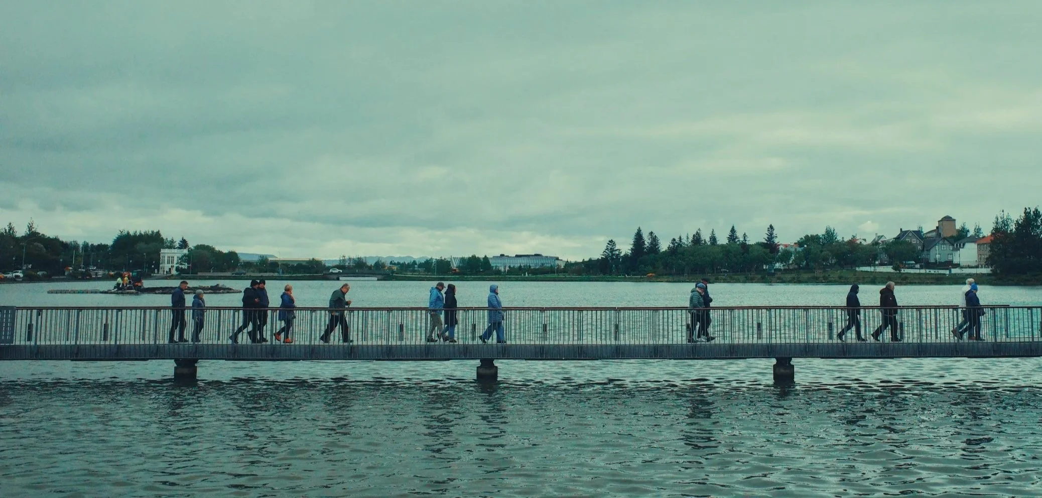 People walking across a suspension bridge over water on a cloudy day.