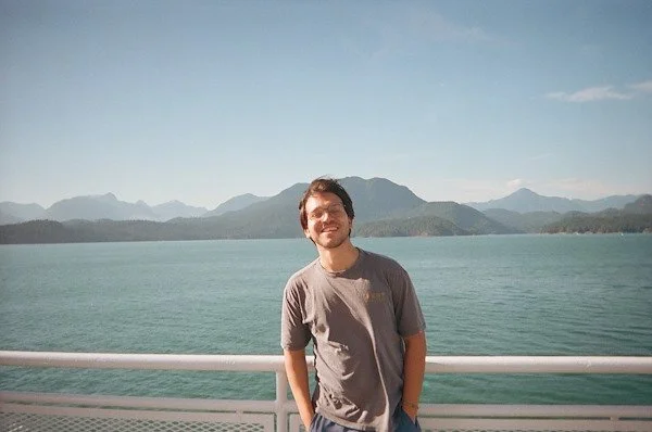 A young man smiles standing on a boat deck with mountains and water in the background.