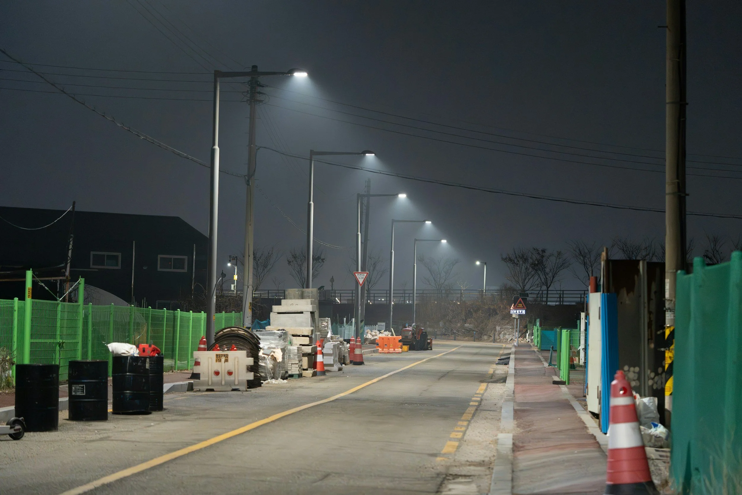 Nighttime scene of a road under construction with streetlights illuminating the area, construction cones and barriers on the side, and construction equipment and materials scattered along the sidewalk.