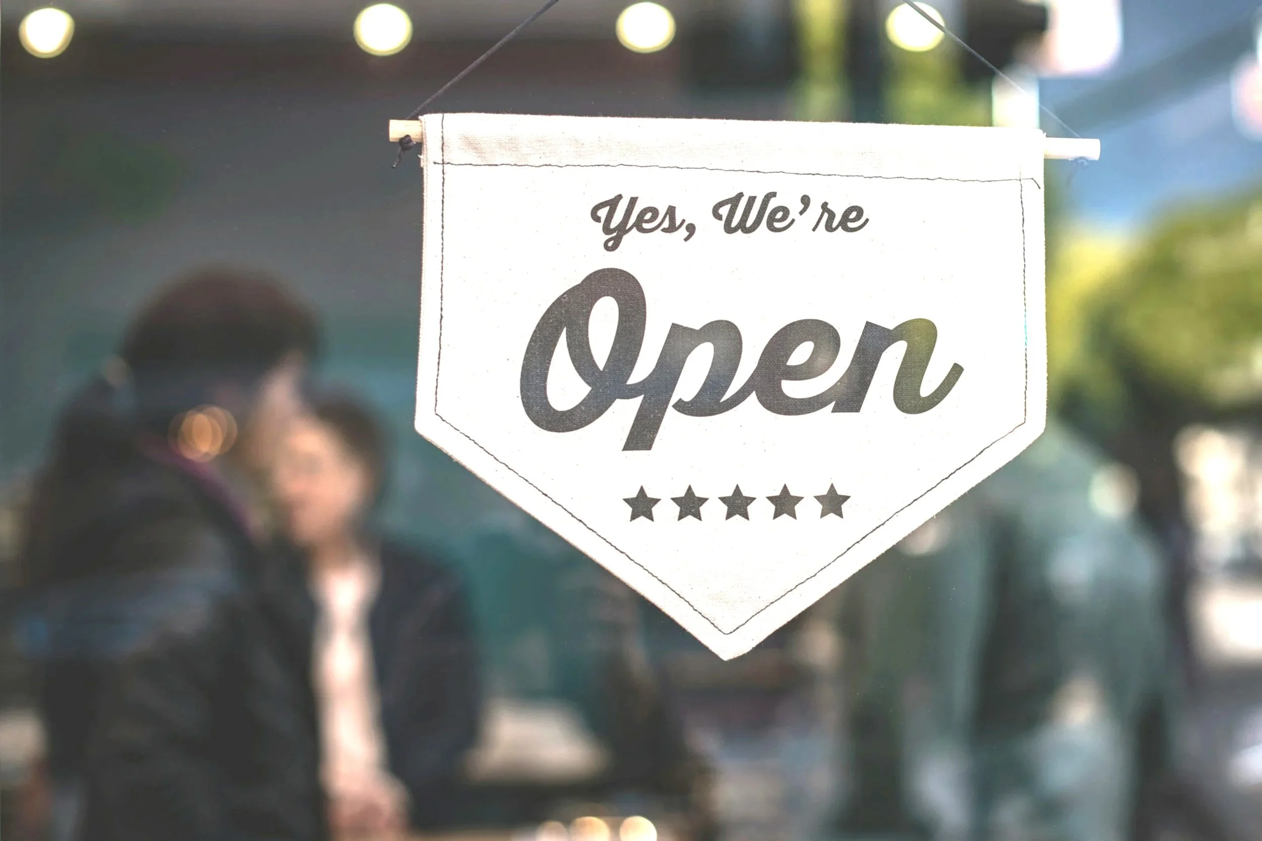 A white sign hanging on a storefront window reading "Yes, We're Open" with the word "Open" in large script and five stars below. Blurred people are visible in the background outside the store.