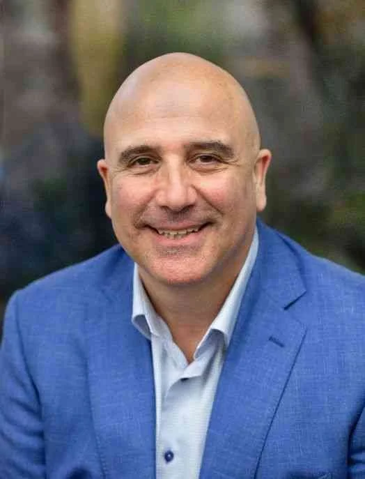 A smiling bald man wearing a blue suit and a white shirt, outdoors with a blurred background of trees.