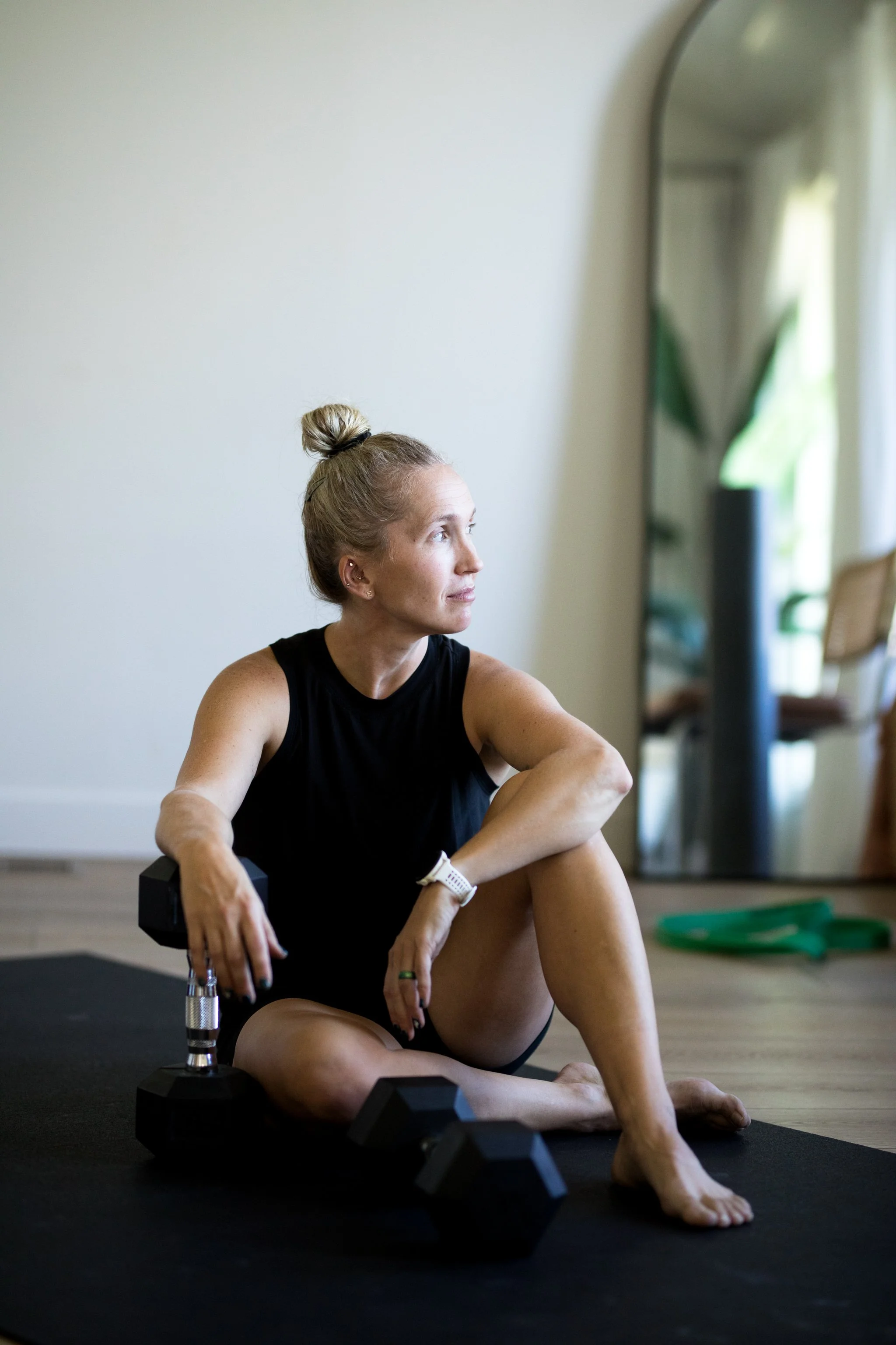 A woman sitting cross-legged on a black exercise mat in a bright room, holding a dumbbell in one hand and looking thoughtfully to her right. There are two more dumbbells and a water bottle near her on the mat, with exercise equipment and plants visible in the background.
