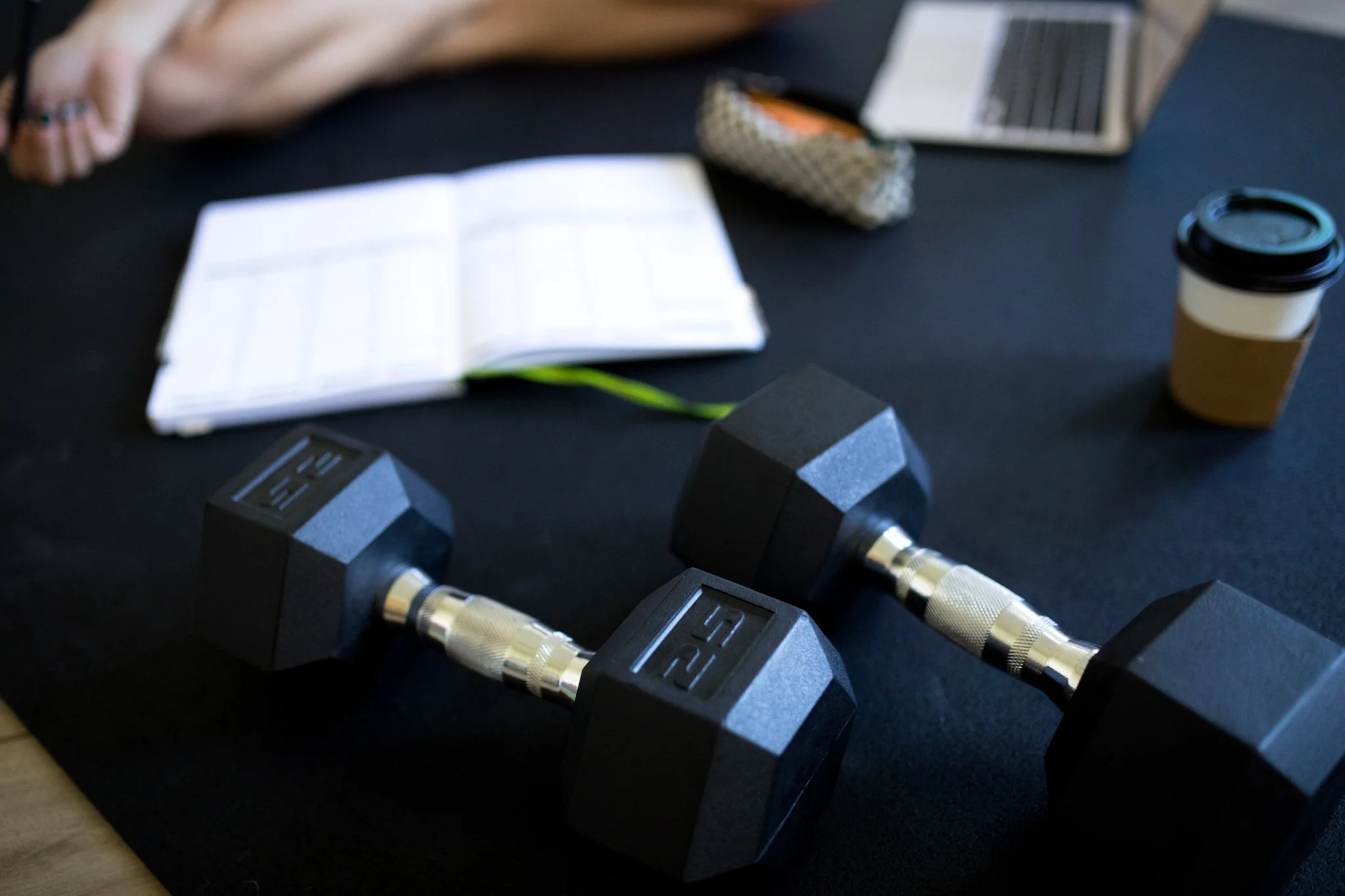 Dumbbells, a notebook, a laptop, a coffee cup, and fun pens on a black exercise mat, indicating a workspace or gym setting.