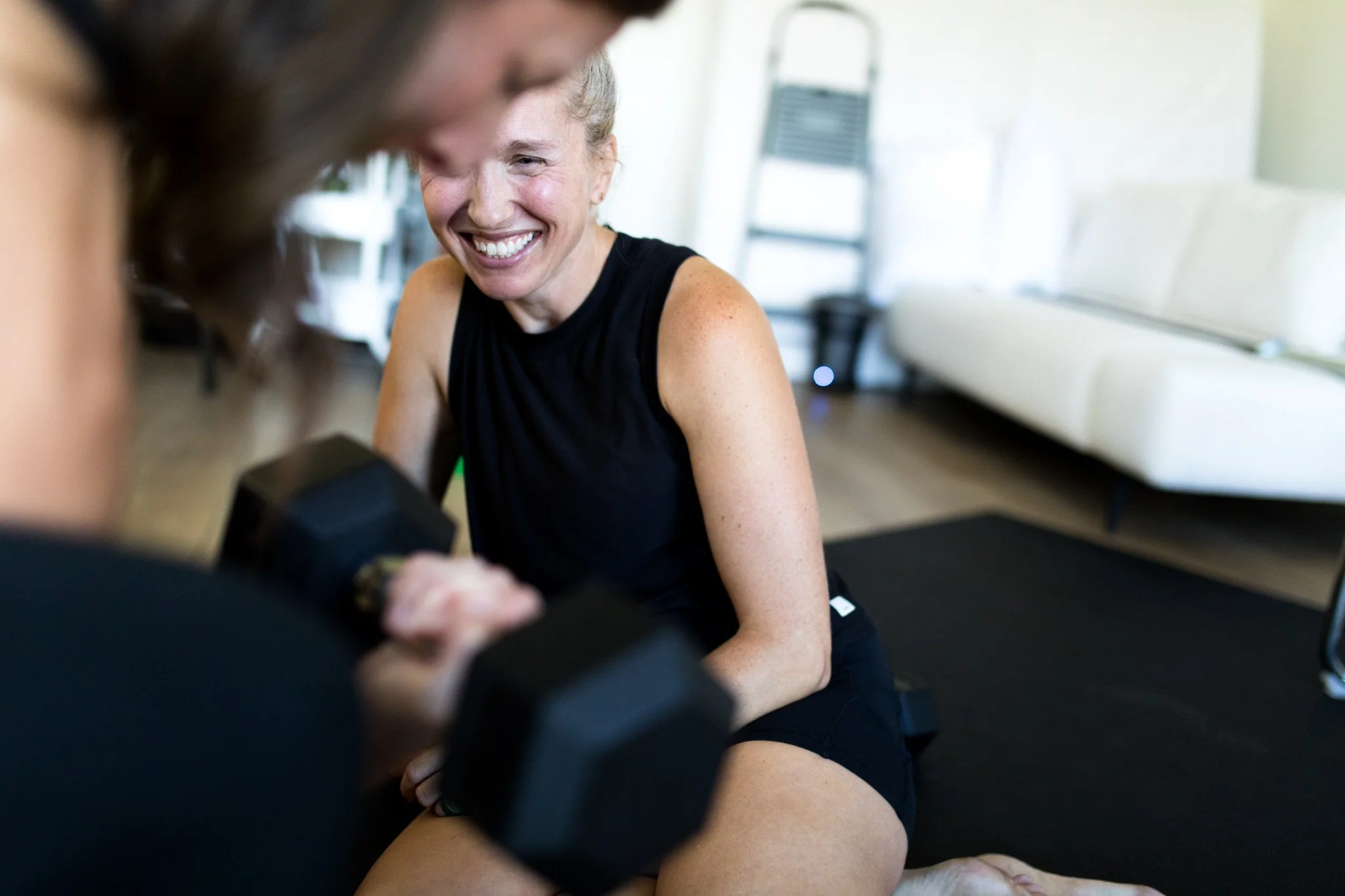 Two women exercising in a bright fitness studio, smiling, with one woman kneeling and the other sitting on the floor near a workout bench.