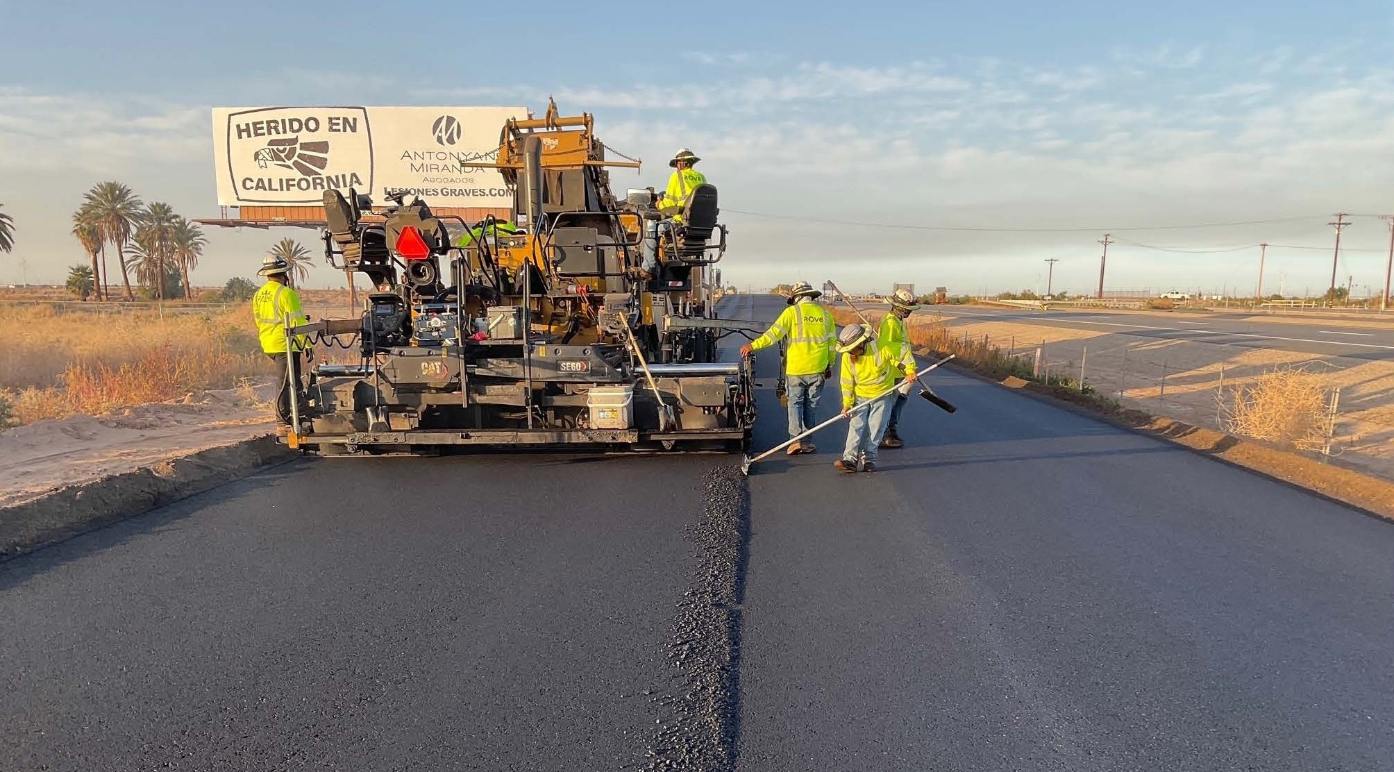 Workers in yellow safety vests and white hats laying down fresh asphalt on a road with a paving machine in a rural area with palm trees in the background.