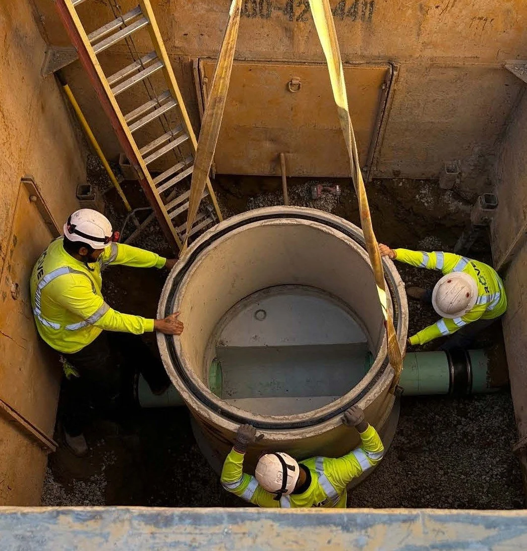 Four construction workers in yellow safety vests and white helmets are installing a large concrete pipe in a construction site. The workers are positioned around the pipe, which is in a square excavation with wooden and metal supports.
