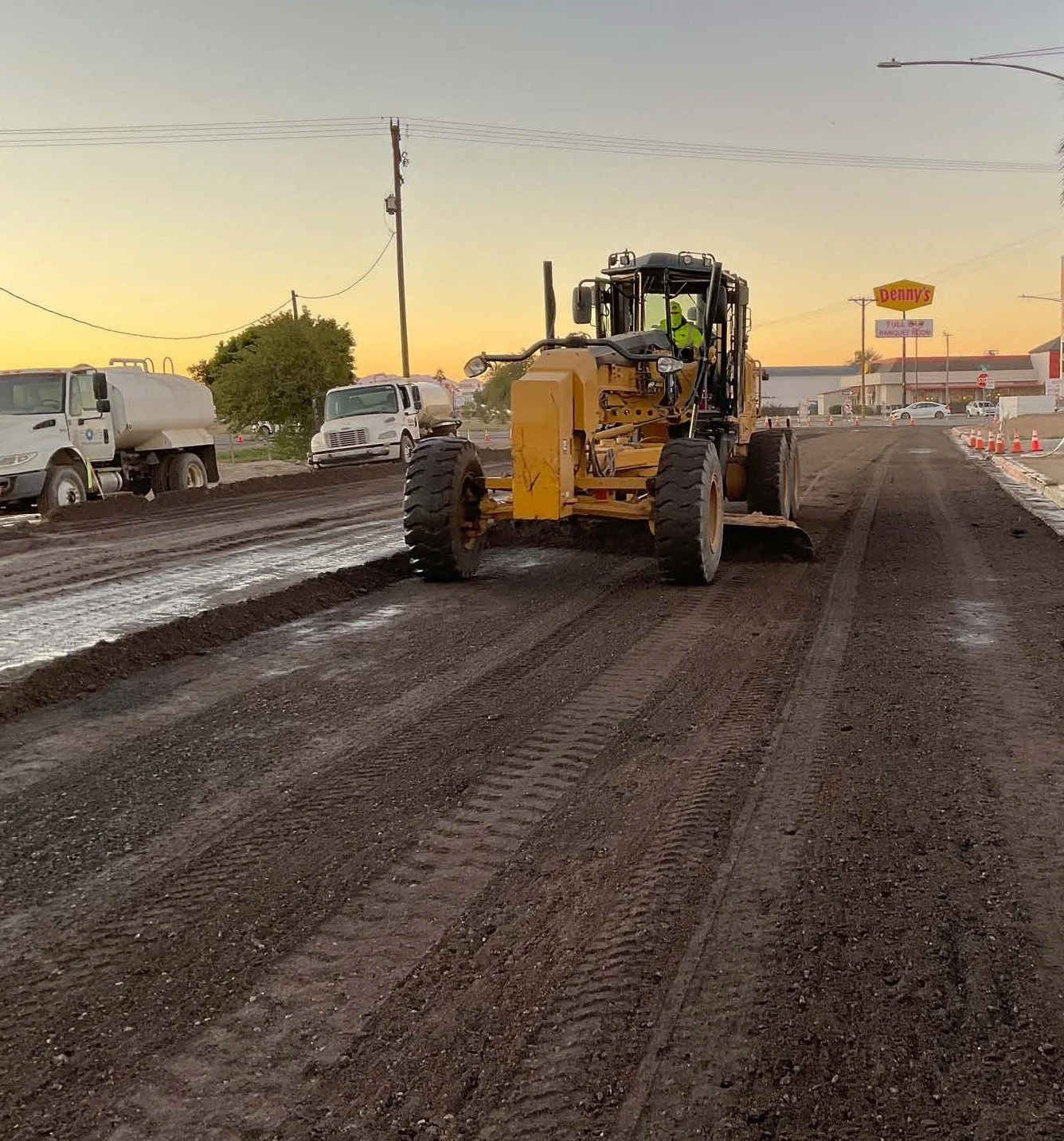 A construction worker operating a yellow road grader smoothing freshly laid asphalt on a street during sunset, with parked trucks and a Denny's restaurant in the background.