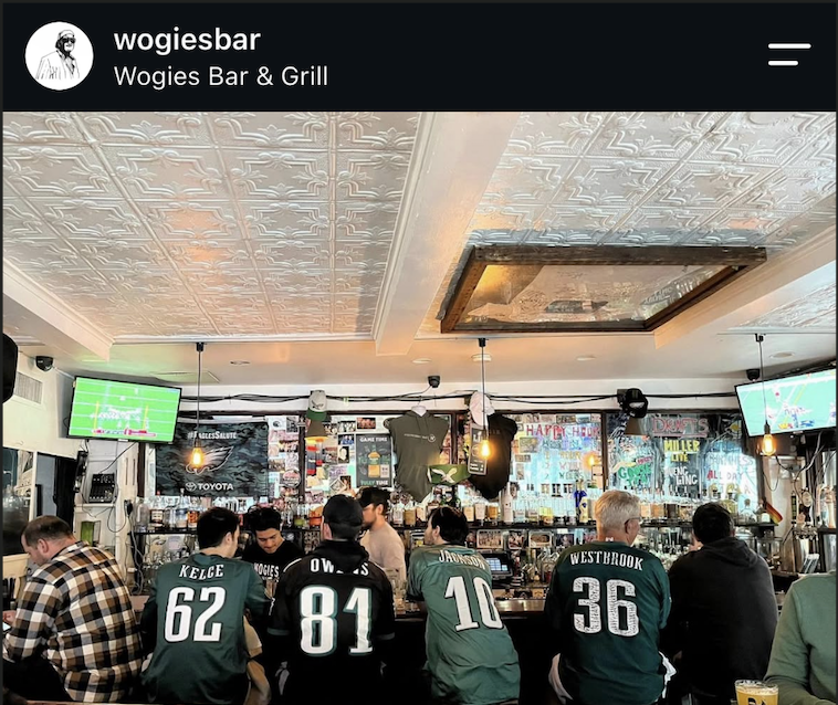 Interior of Wogies West Village during an Eagles football game. Fans wearing Philadelphia Eagles jerseys sit along a crowded bar, watching football on TVs. The restaurant features a decorative tin ceiling, hanging lights, and sports memorabilia.