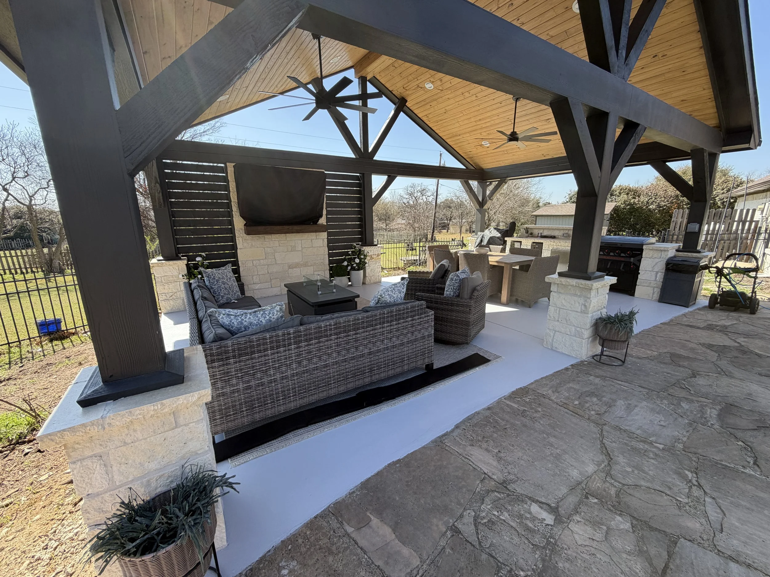Backyard patio area under a wooden overhang with ceiling fans, wicker furniture, a stone fireplace, and a gas grill. There are potted plants and a view of trees and neighboring houses in the background.