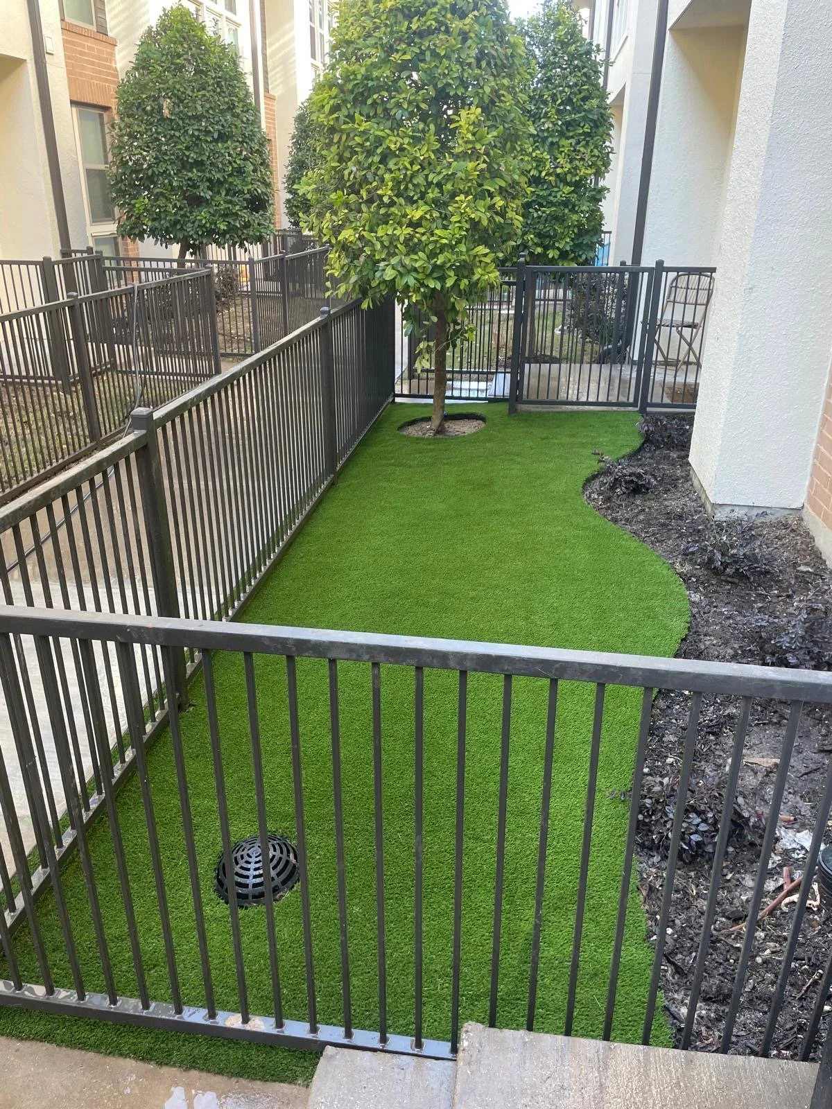 Small backyard with artificial grass, a mature tree in the center, enclosed by metal fencing, with a small garden bed on the right and a patio area in the background.