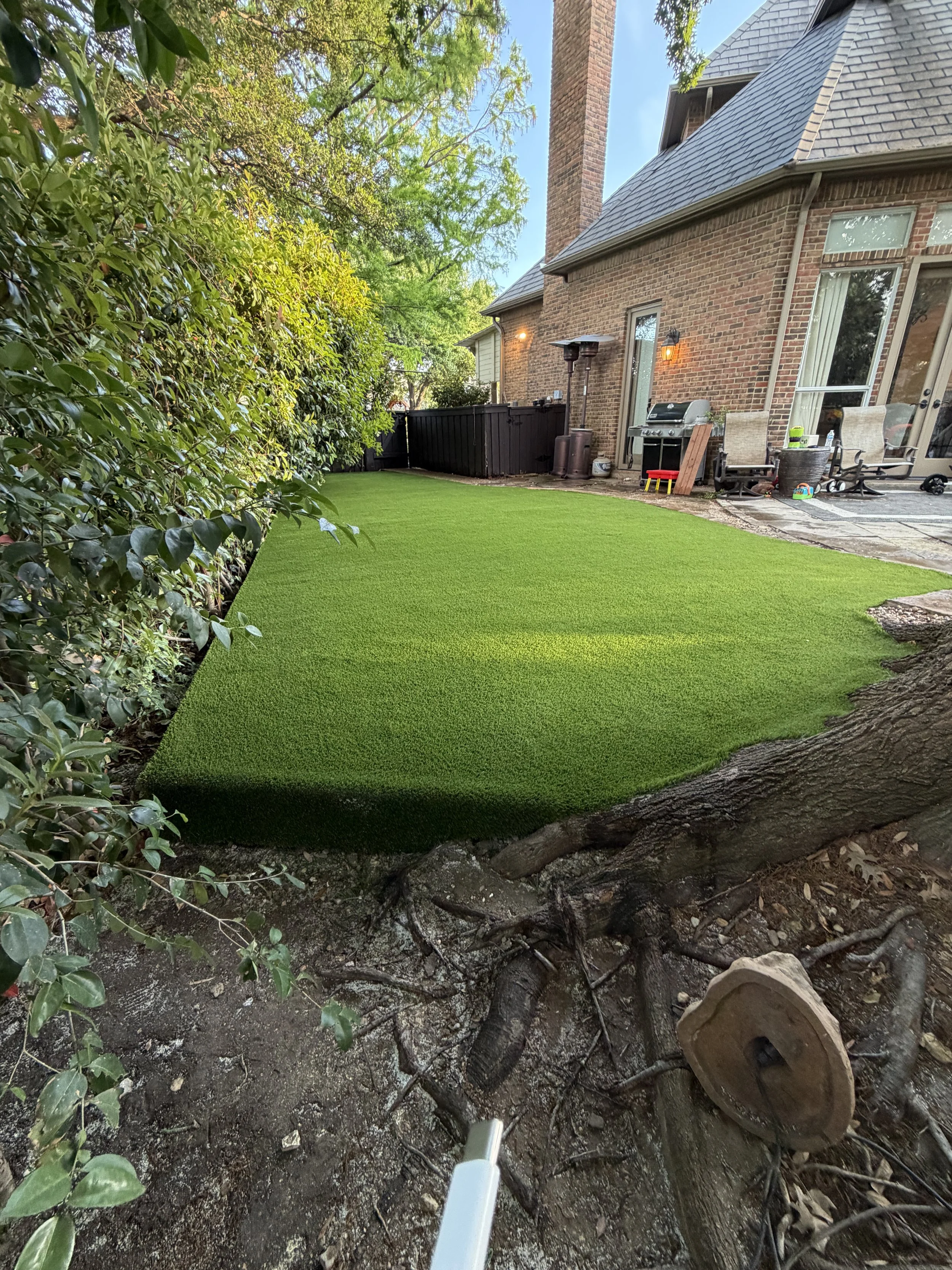 Backyard with a green artificial lawn, a brick house with a chimney, some patio furniture, a barbecue grill, and a large tree in the foreground with exposed roots.