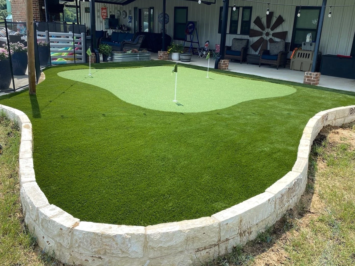 Backyard putting green with a flag, surrounded by a stone border, under a covered patio with outdoor furniture and decorative items.