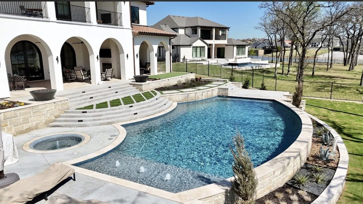 Exterior view of a luxurious backyard with a large kidney-shaped swimming pool, a hot tub, and a spacious patio area with lounge chairs, surrounded by a manicured lawn and neighboring houses.