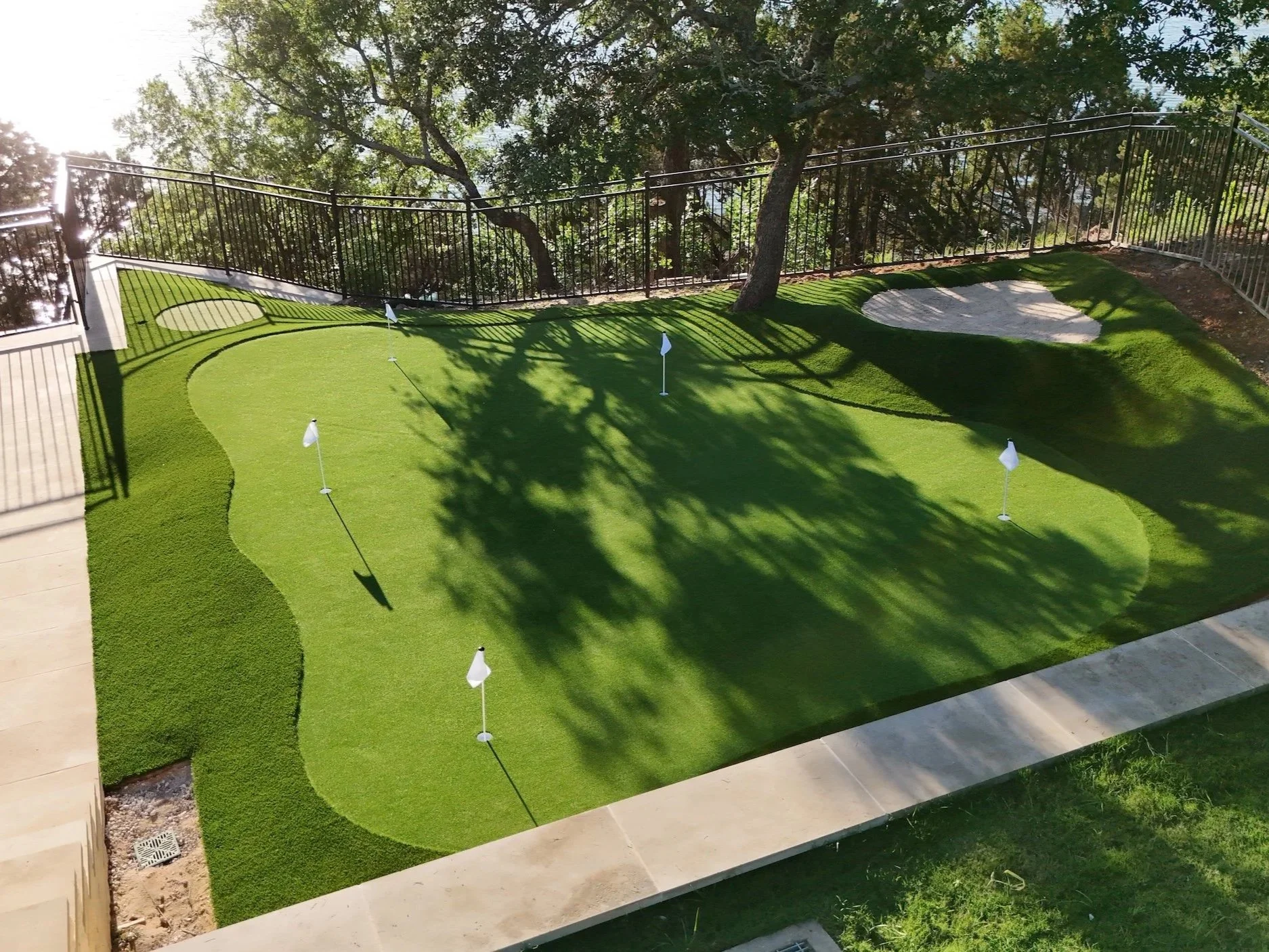 A backyard putting green with a grass surface, small flags, a sand bunker, trees in the background, and a black metal fence surrounding the area.