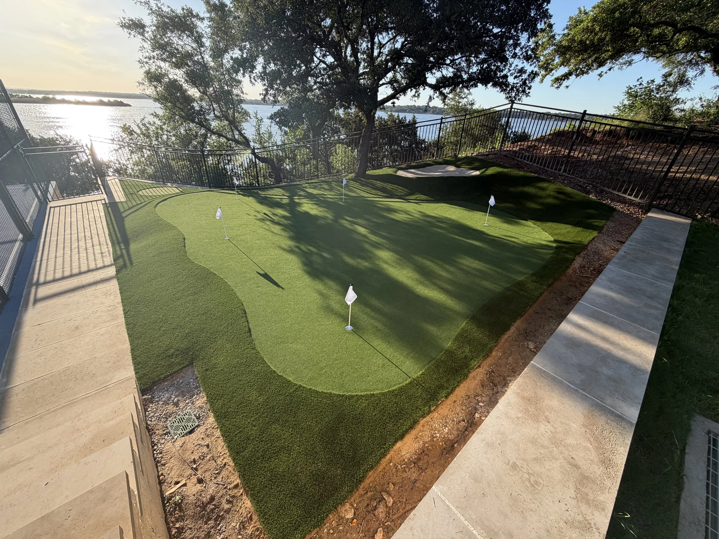 A backyard with a small putting green near a lake, surrounded by trees and a black metal fence, with a concrete pathway and a patch of dirt.