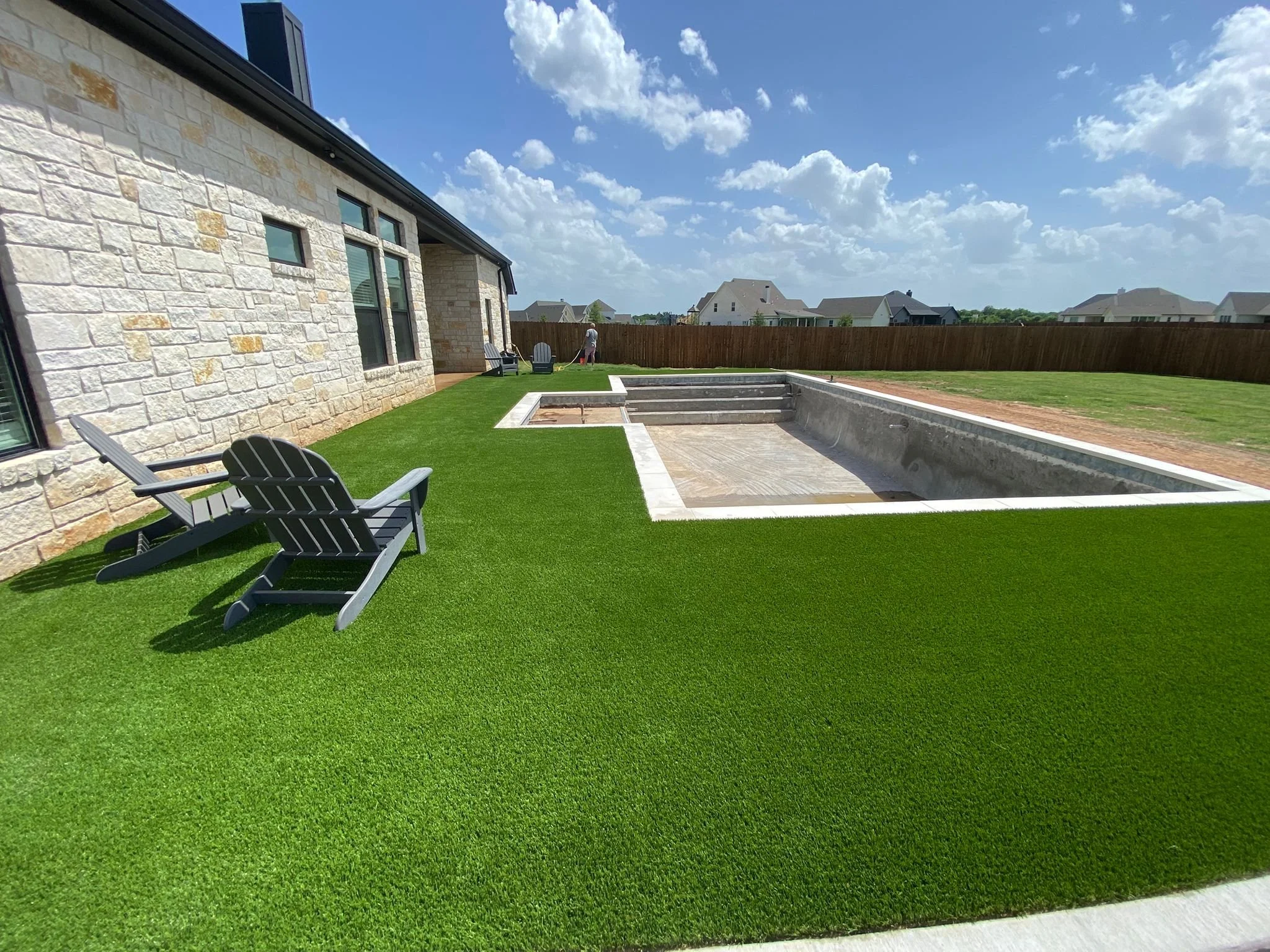 Backyard with artificial grass, chairs, a pool under construction, and a person watering the grass, with a wooden fence and neighboring houses in the background.