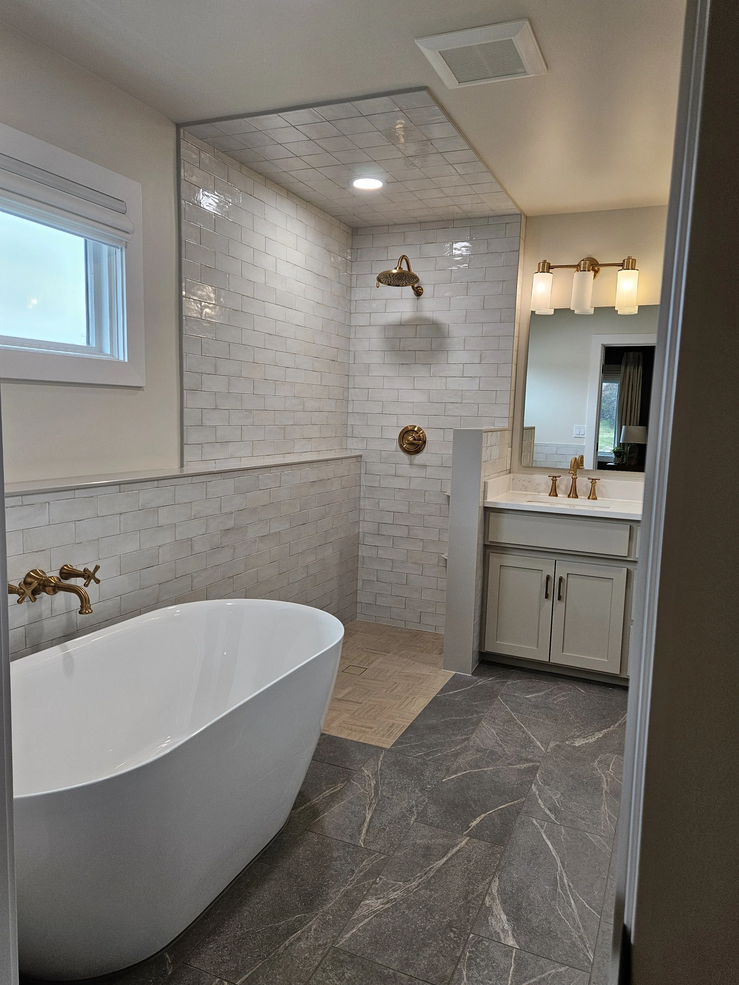 Modern bathroom with a freestanding bathtub, white tiled shower area with brass fixtures, a vanity with a mirror and three light fixtures, gray marble floor, and a window with white trim.