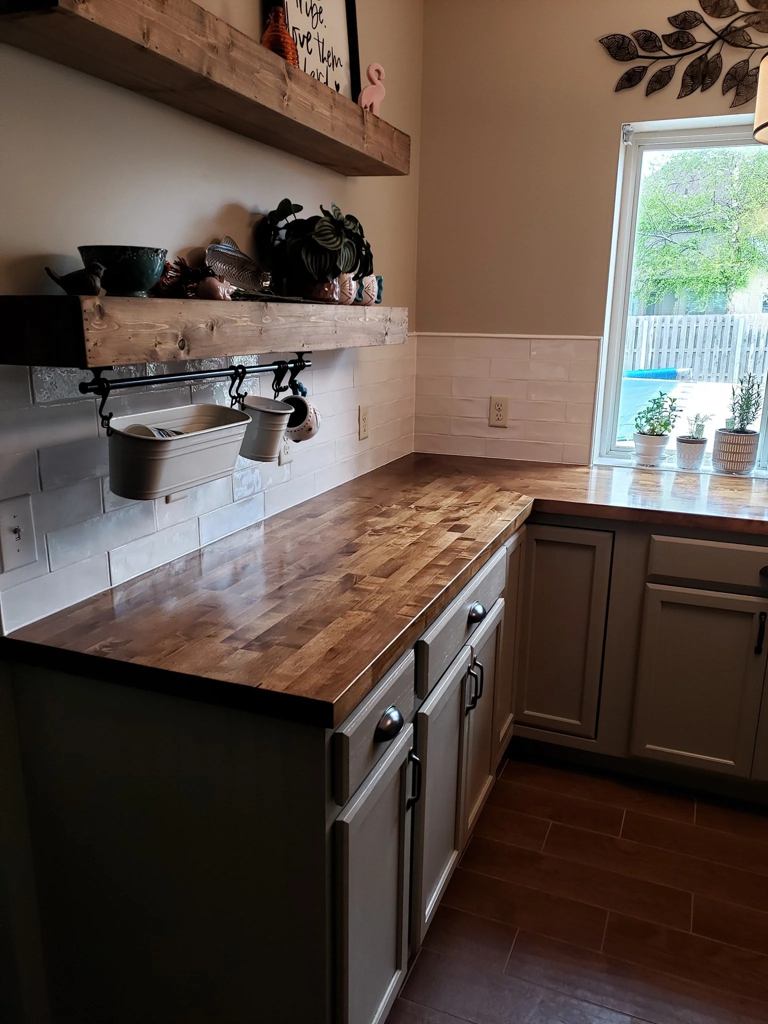 Kitchen with wooden countertop, white cabinets, and a window with a view of trees outside. Shelves hold kitchen decor, plants, and utensils.