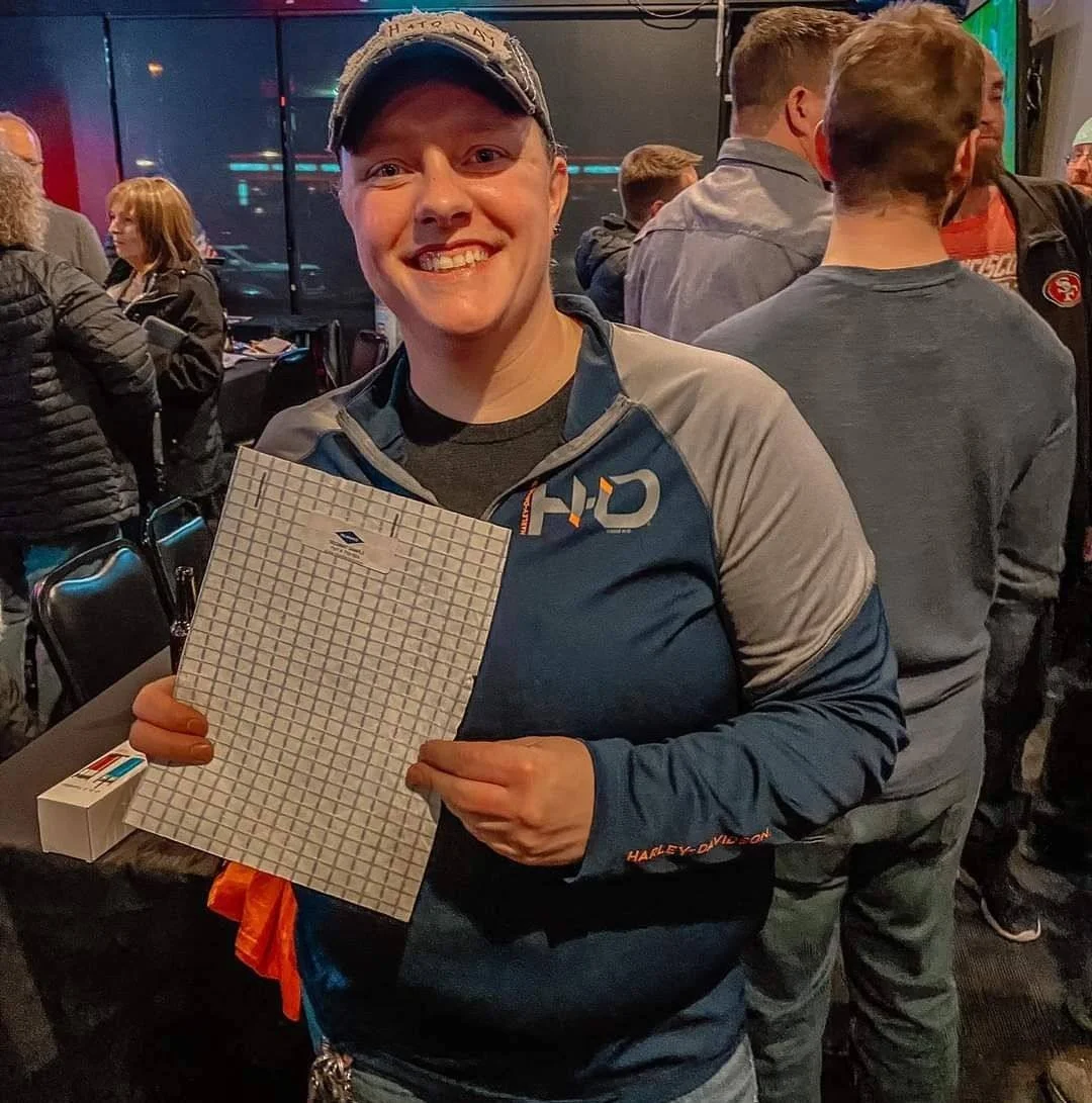 A smiling woman in a blue and gray Harley-Davidson jacket holding a graph paper and a small box, in a crowded indoor setting with people in the background.