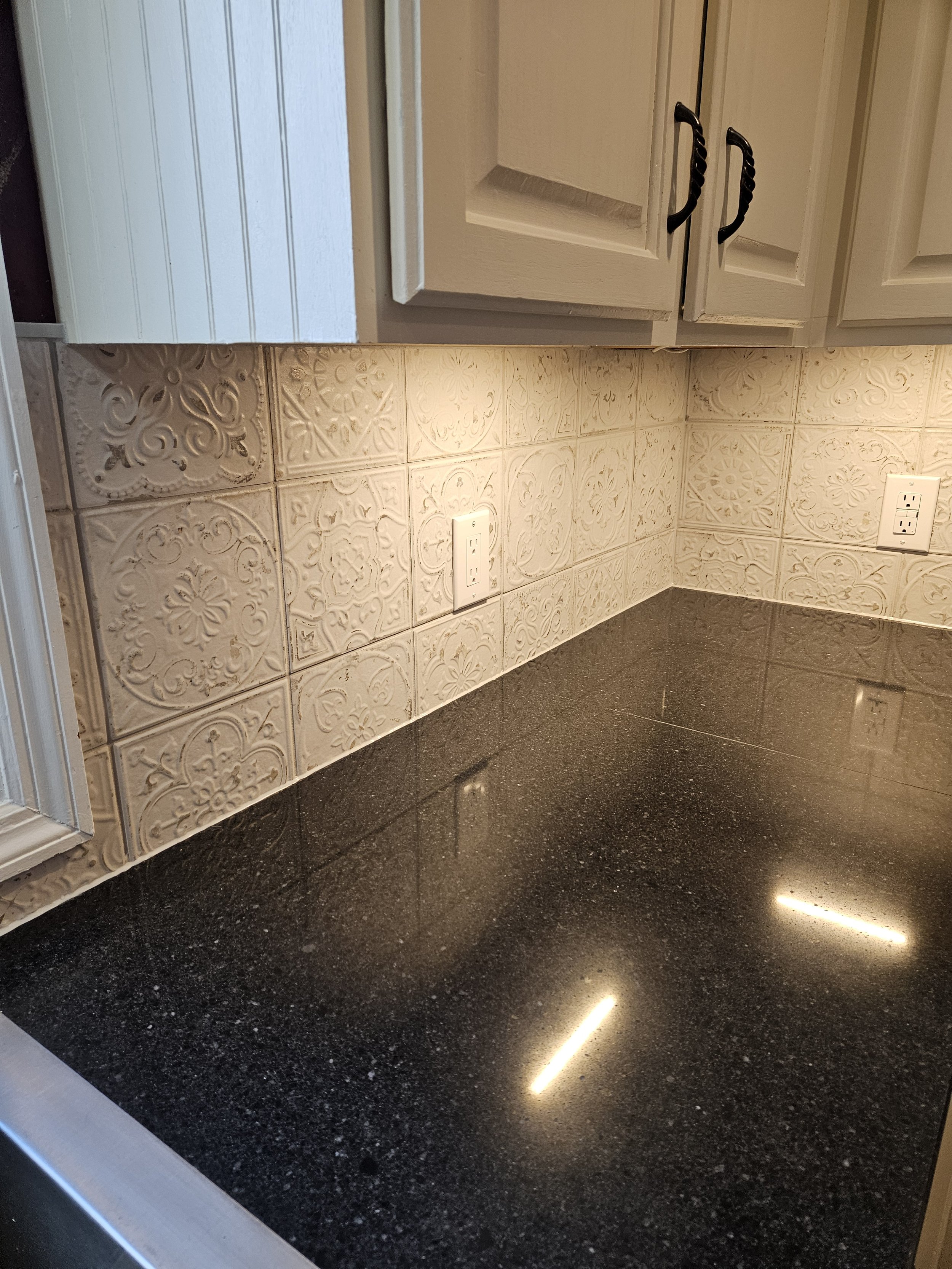 A kitchen countertop with decorative beige tiles and two electrical outlets, with dark granite surface and a wooden cabinet above.