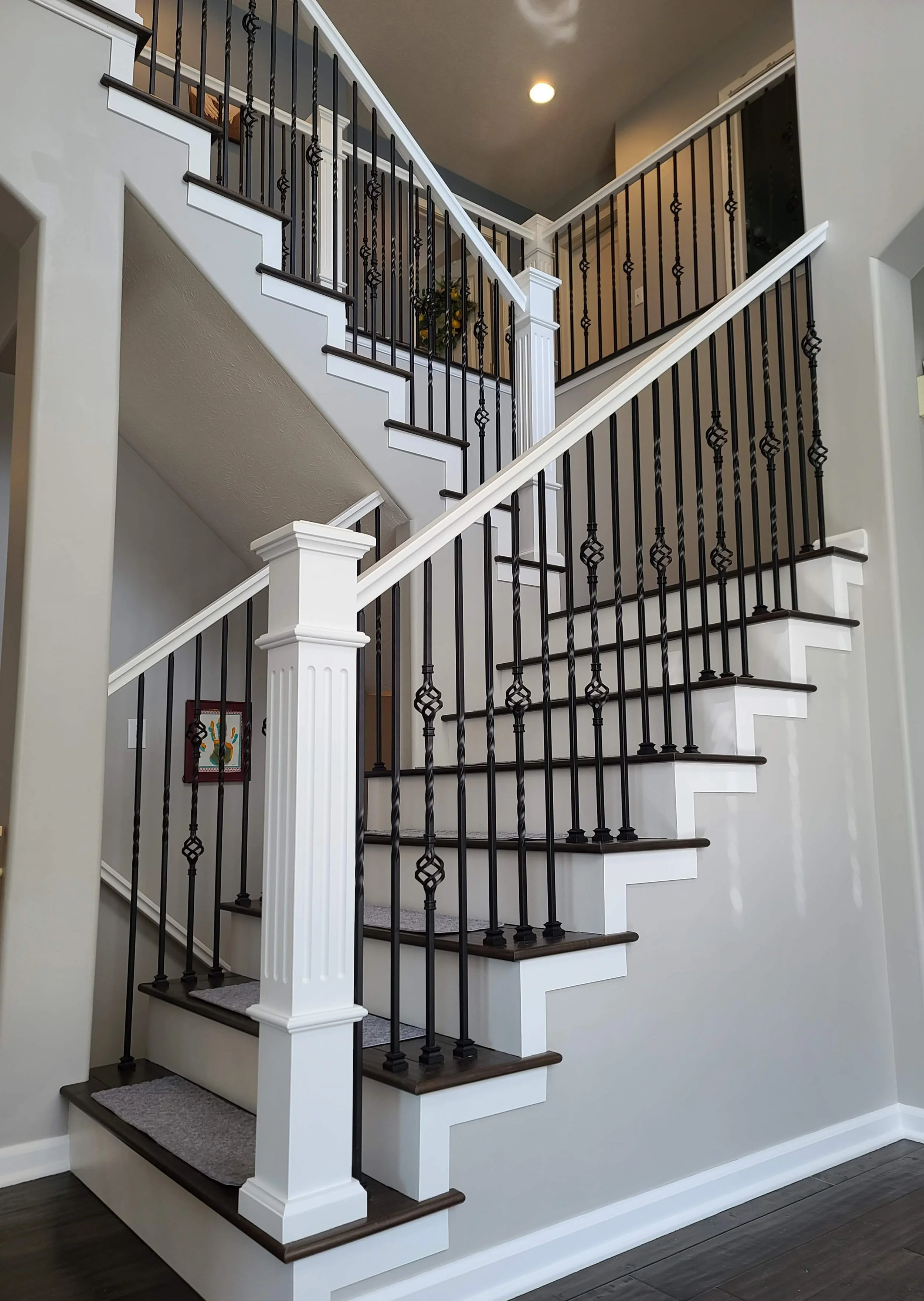 Interior view of a staircase with white risers, dark wooden treads, decorative black metal balusters, and white newel posts, with gray walls and dark flooring.