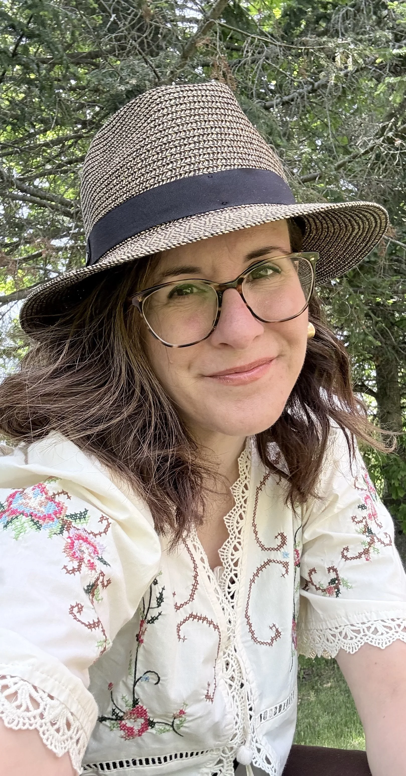 Une femme souriante portant un chapeau en paille à bande noire, des lunettes et une chemise blanche brodée de motifs floraux, dans un environnement verdoyant.