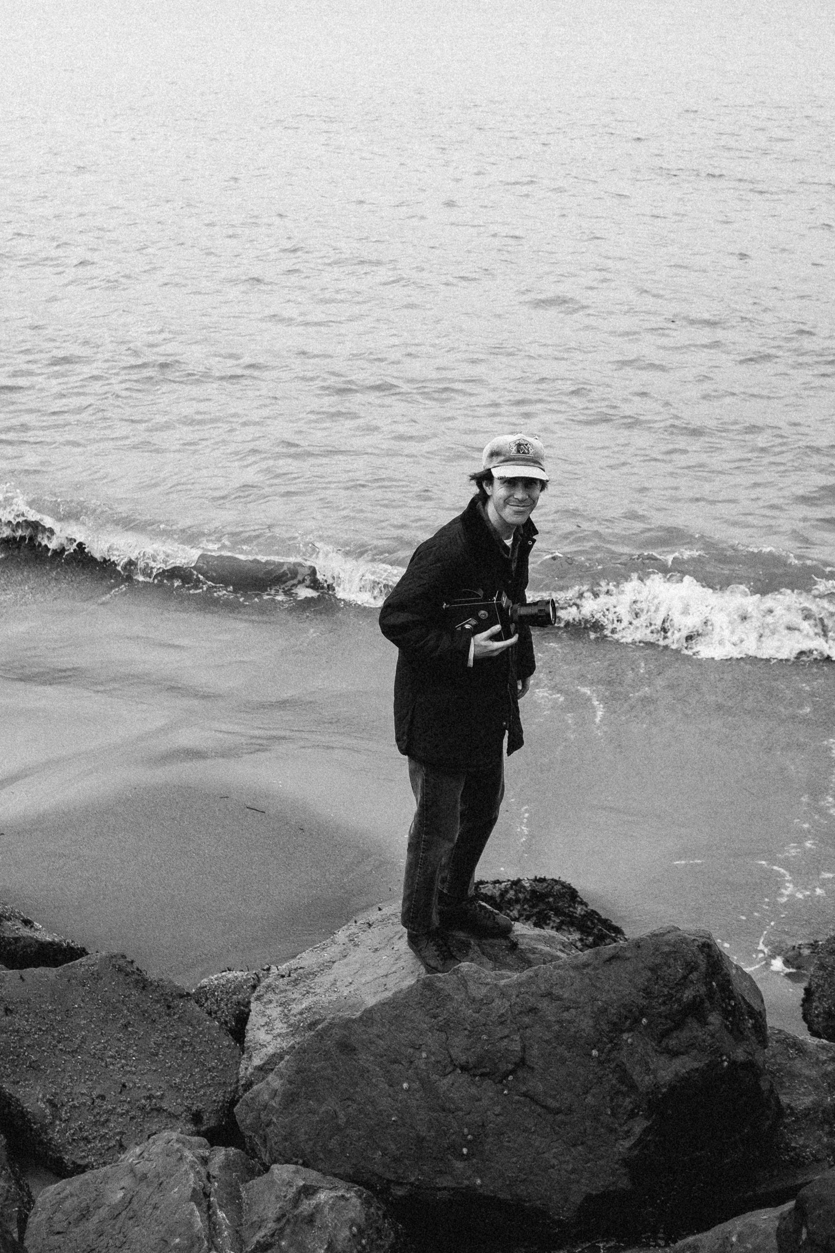 A man stands on rocks by the water's edge, holding a 16mm camera, with gentle waves in the background.