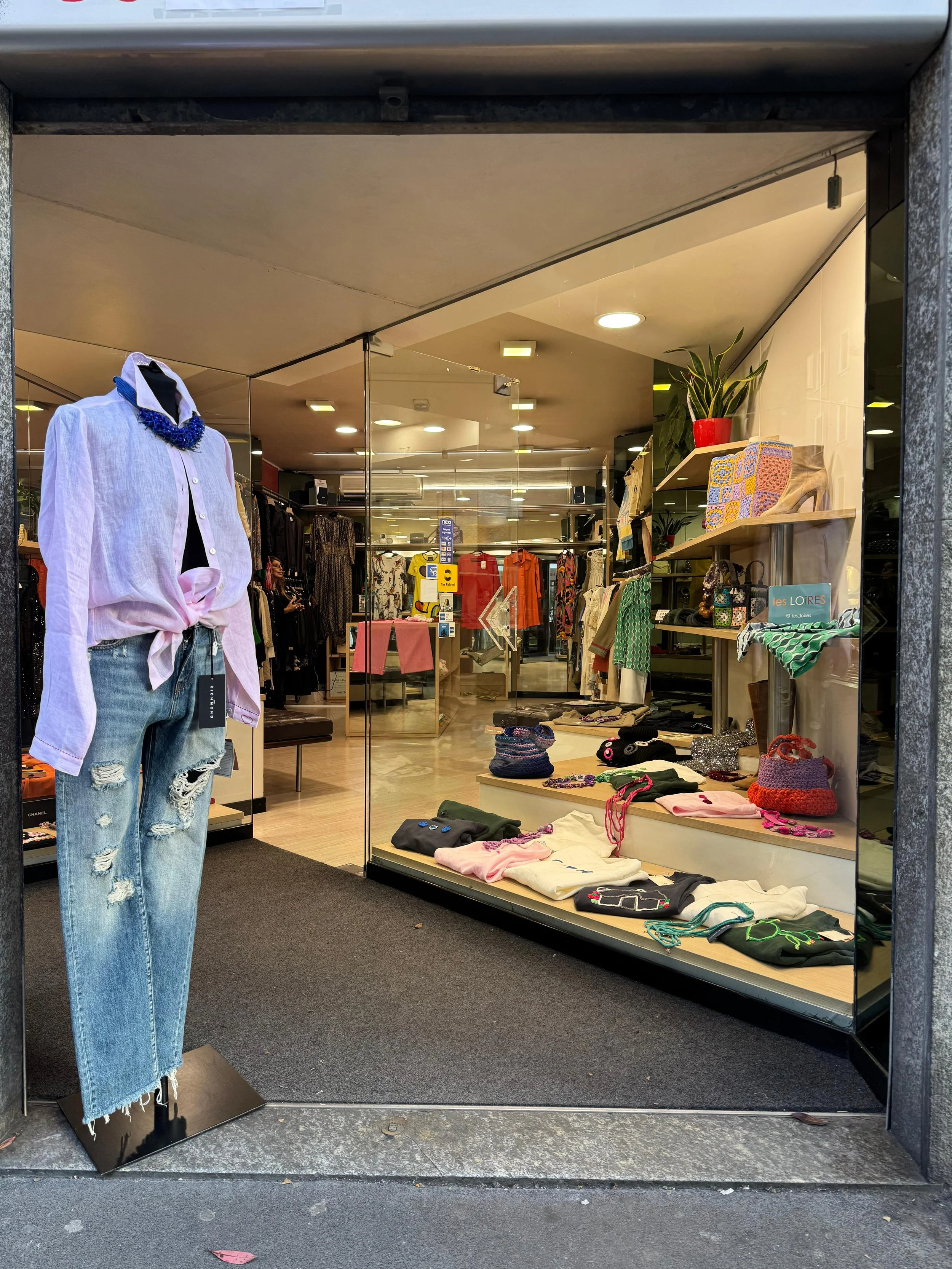 Clothing store display window with a mannequin dressed in a white shirt, distressed jeans, and a blue necklace, and shelves with folded clothes, bags, and accessories inside the store.