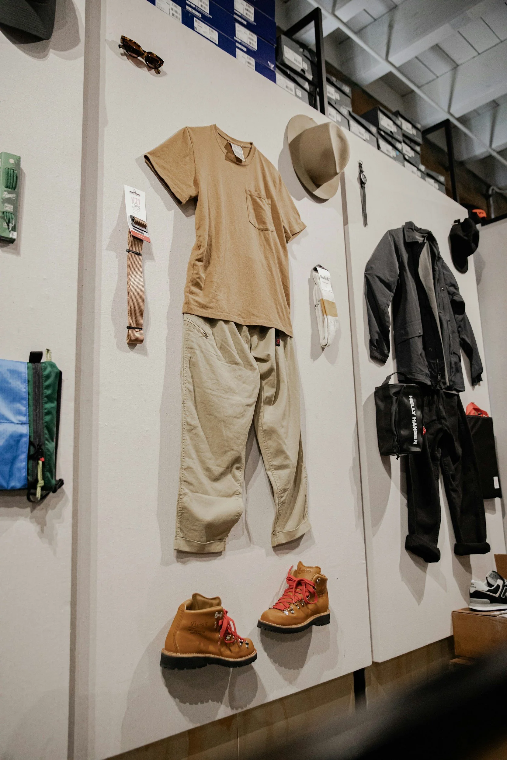 Display of outdoor gear including a beige t-shirt and pants, a hat, sunglasses, a watch, and hiking boots on a retail wall.