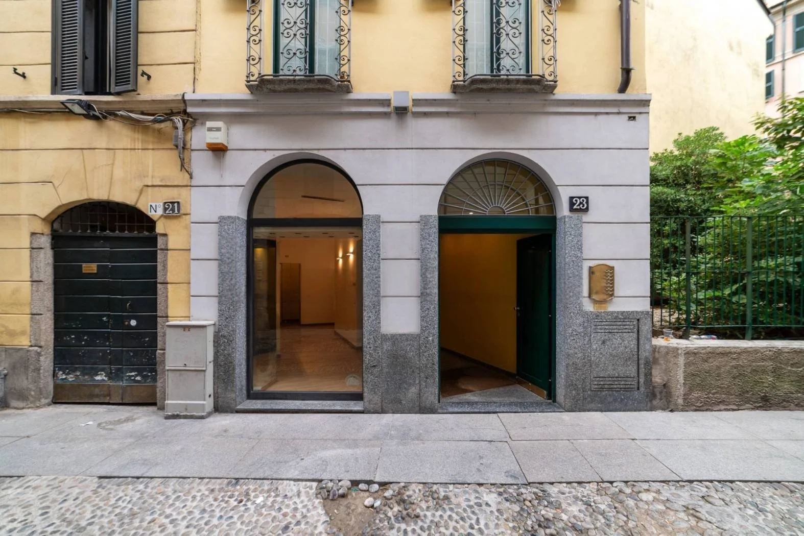 Facade of a building with two entrances, one glass door and one open archway, with apartment numbers 21 and 23, and decorative metal balconies above. To the left, a garage door, and to the right, a mailbox and a green fence with greenery behind.
