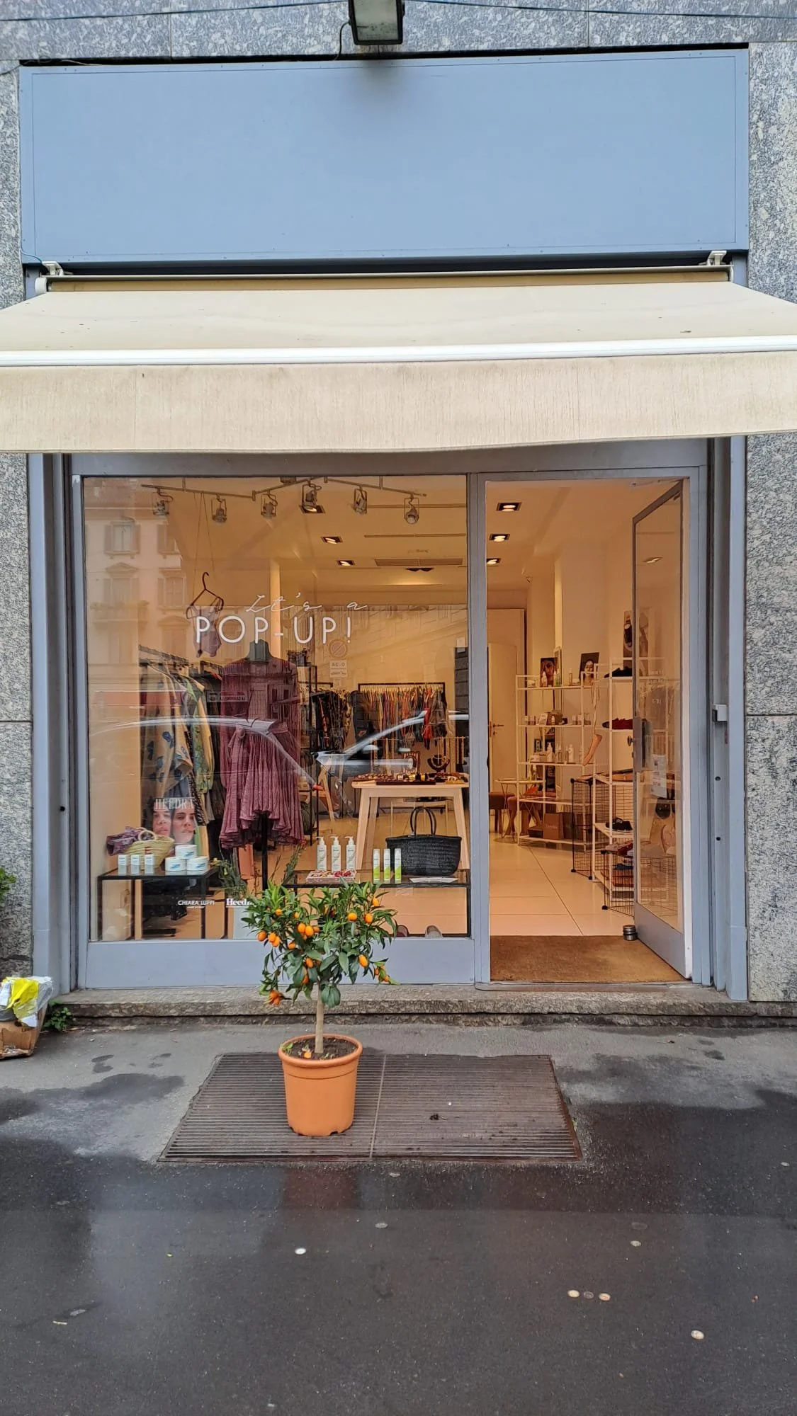 The storefront of a clothing boutique with large glass windows and an open door, displaying racks of clothes inside. A potted citrus tree with ripe oranges stands outside on a sidewalk, with a doormat in front of the entrance. The store sign indicates a pop-up event.