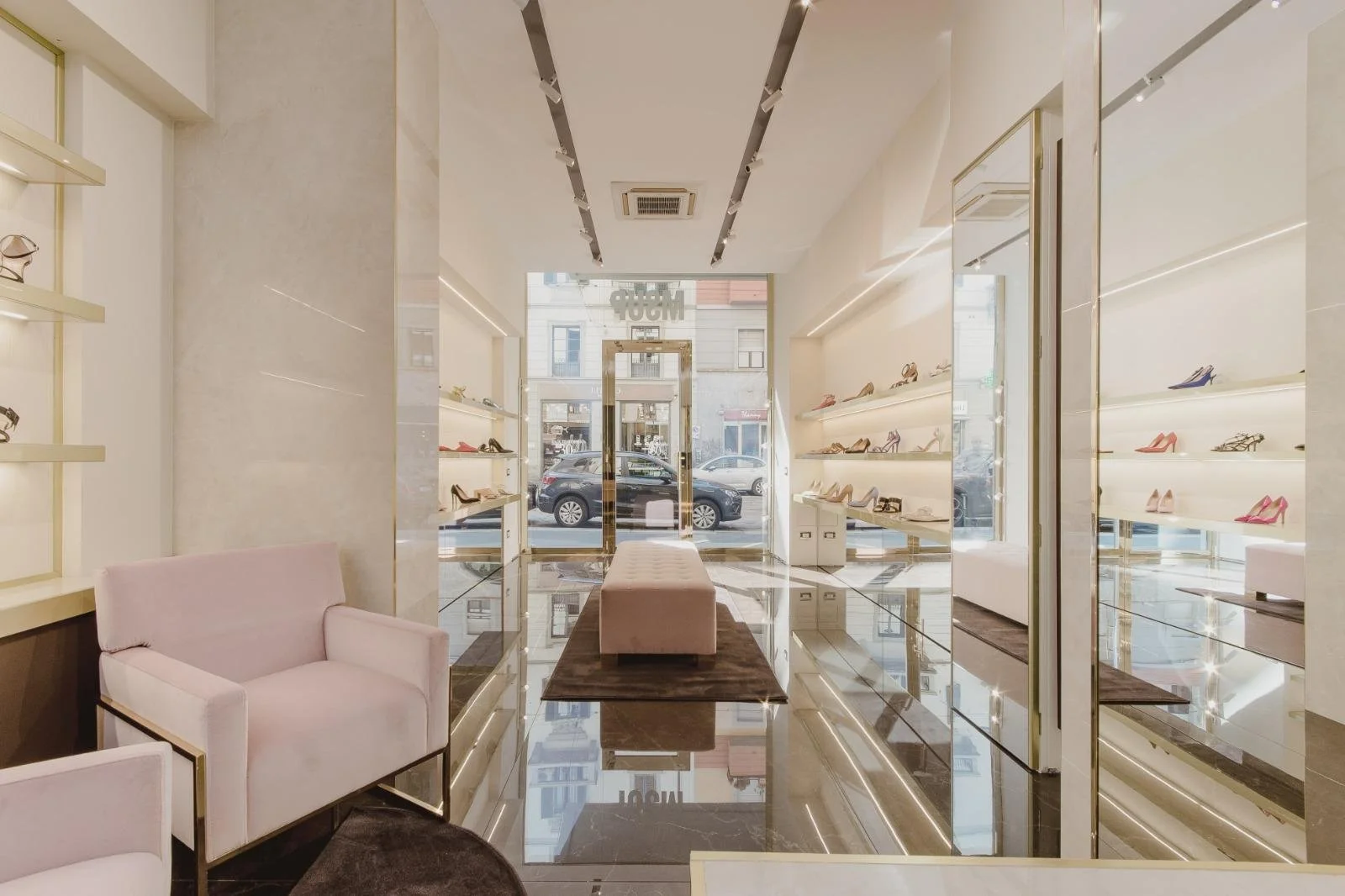 A modern shopping boutique with pink chairs, glass shelves displaying high-heeled shoes, and a pink seating bench in front of large glass windows showing parked cars and a city street outside.