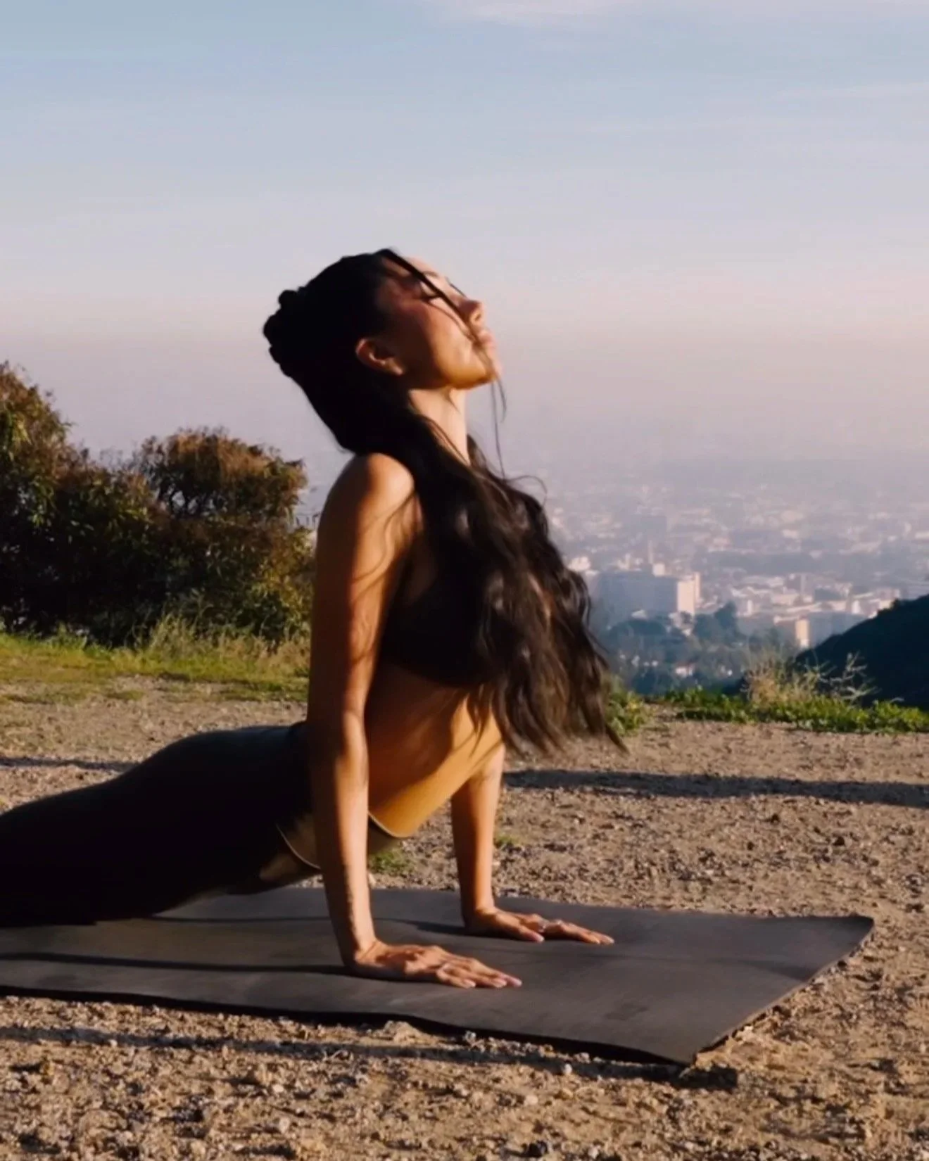 A woman practicing yoga outdoors on a mat with a cityscape and mountainous landscape in the background.